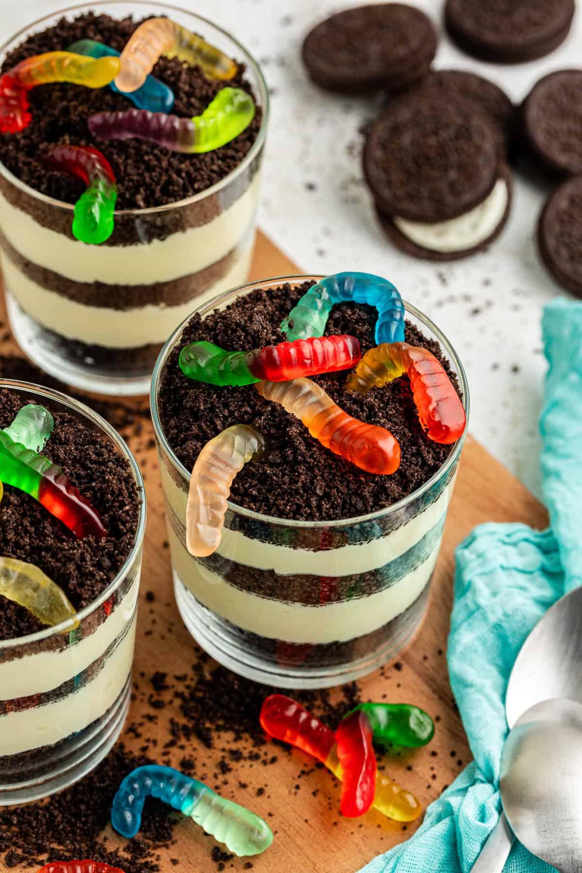 La migliore ricetta per la Torta al Fango 15 Tre piccole coppe da trifle di torta di terra guarnite con vermi gommosi su un piatto di legno circondate da Oreo interi, briciole di Oreo, vermi gommosi, cucchiai e un asciugamano color ottanio chiaro