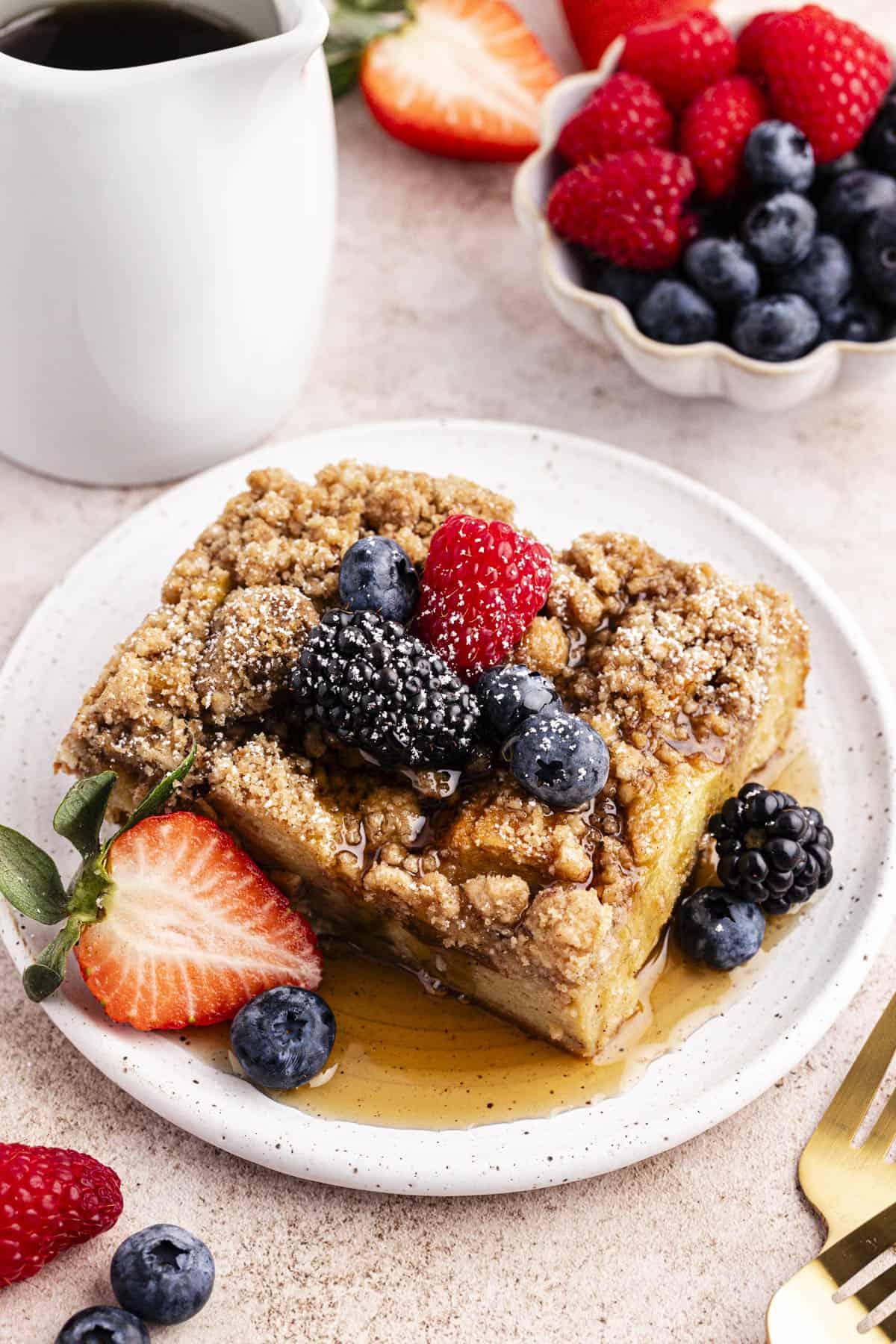 Migliore Ricetta per Budino di French Toast 15 Un piattino con una fetta di casseruola di French Toast guarnita con sciroppo d'acero, frutti di bosco freschi e una leggera spolverata di zucchero a velo accanto a una piccola ciotola di frutti di bosco freschi, un piatto bianco di sciroppo, due forchette e altri frutti di bosco sparsi intorno.