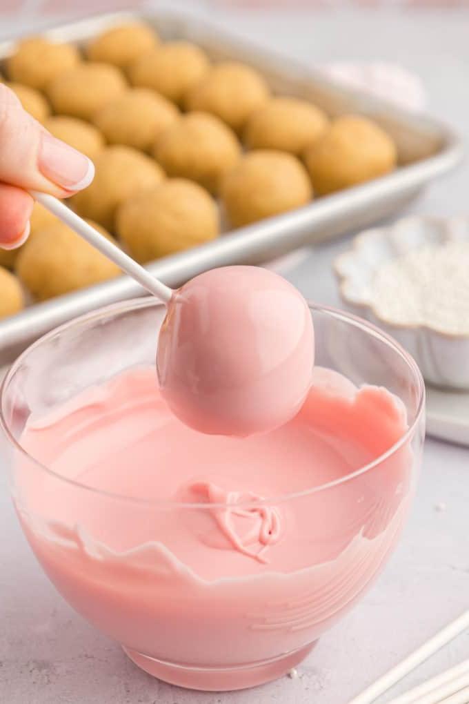un cake pop rosa appena intinto sopra una ciotola di vetro trasparente piena di cioccolato fuso rosa con una teglia di palline di torta semplici sullo sfondo