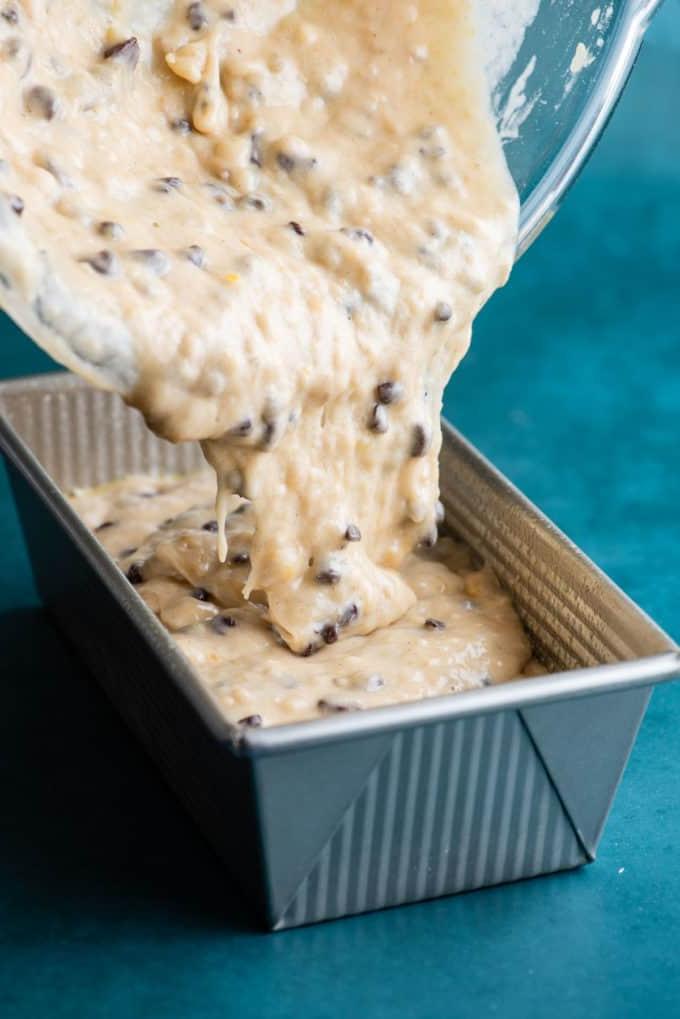 Il Miglior Pan di Banane con Gocce di Cioccolato 15 Impasto del pane alle banane e gocce di cioccolato che viene versato in uno stampo da plumcake su una superficie color ottanio scuro.