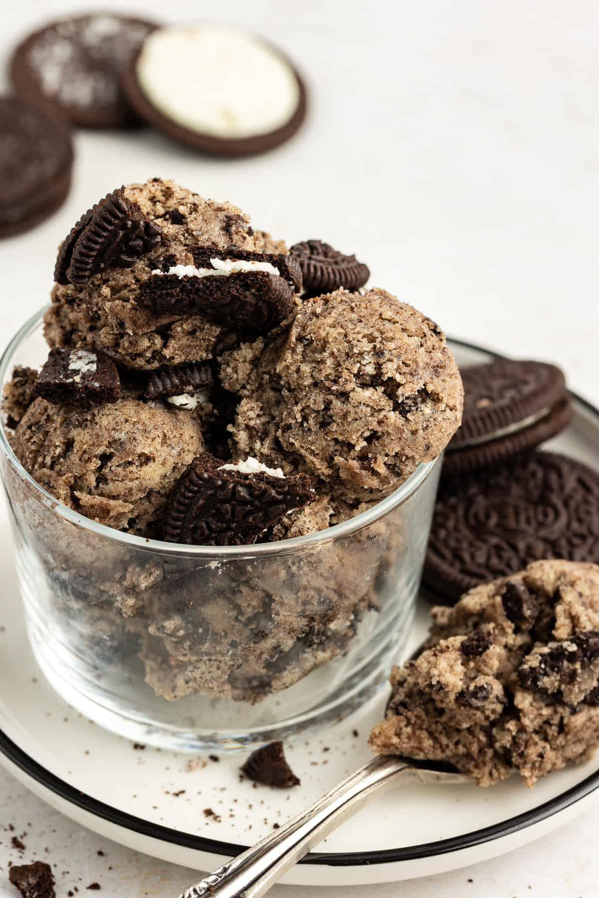 **Semplice Impasto per Biscotti Commestibile** 15 oreo cookie dough balls in a clear glass container sitting on a white plate with a spoonful of edible oreo cookie dough and two whole oreos, plus more oreos scattered in the background