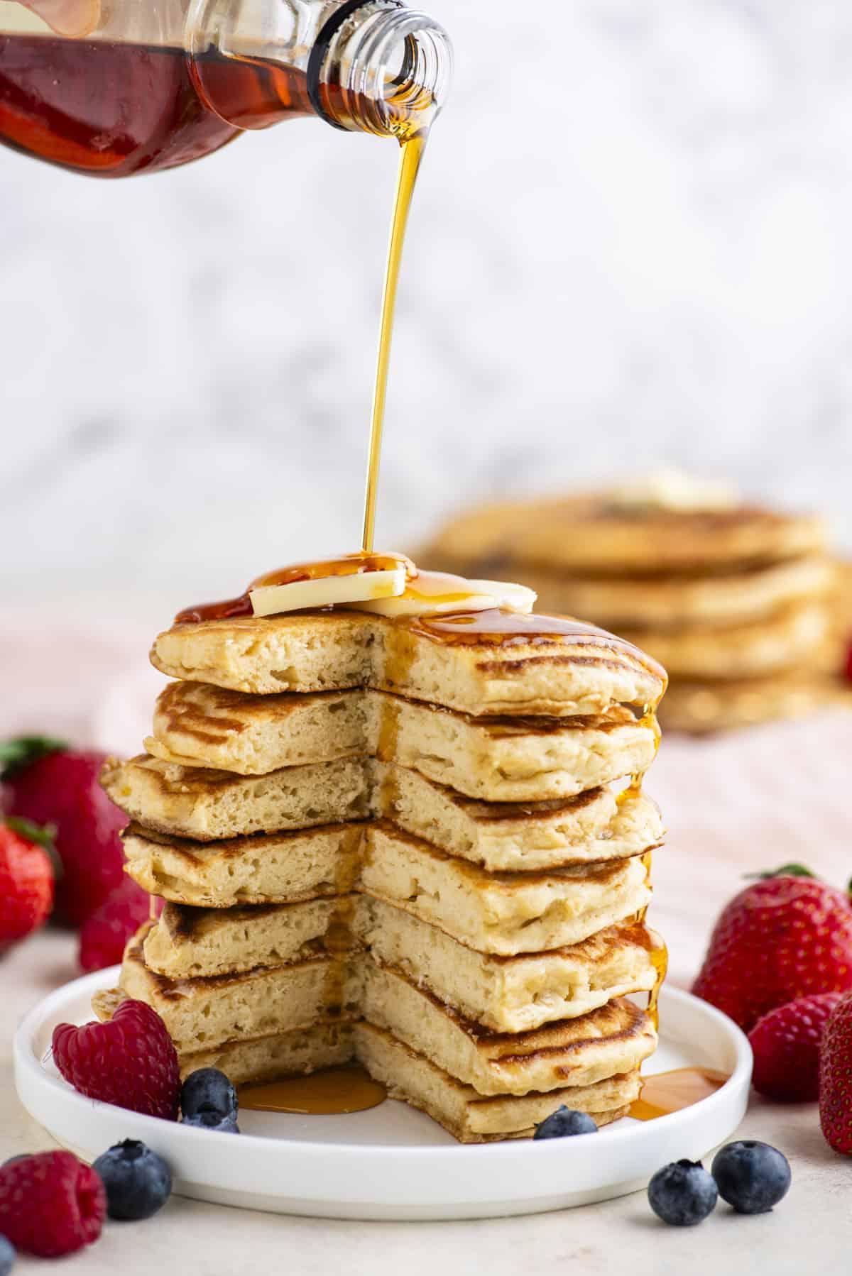 Pancakes Soffici Fatti in Casa 15 Una torre alta di pancake con una porzione triangolare tagliata, condita con sciroppo, sormontata da una fetta di burro e circondata da frutti di bosco freschi su un piatto bianco.