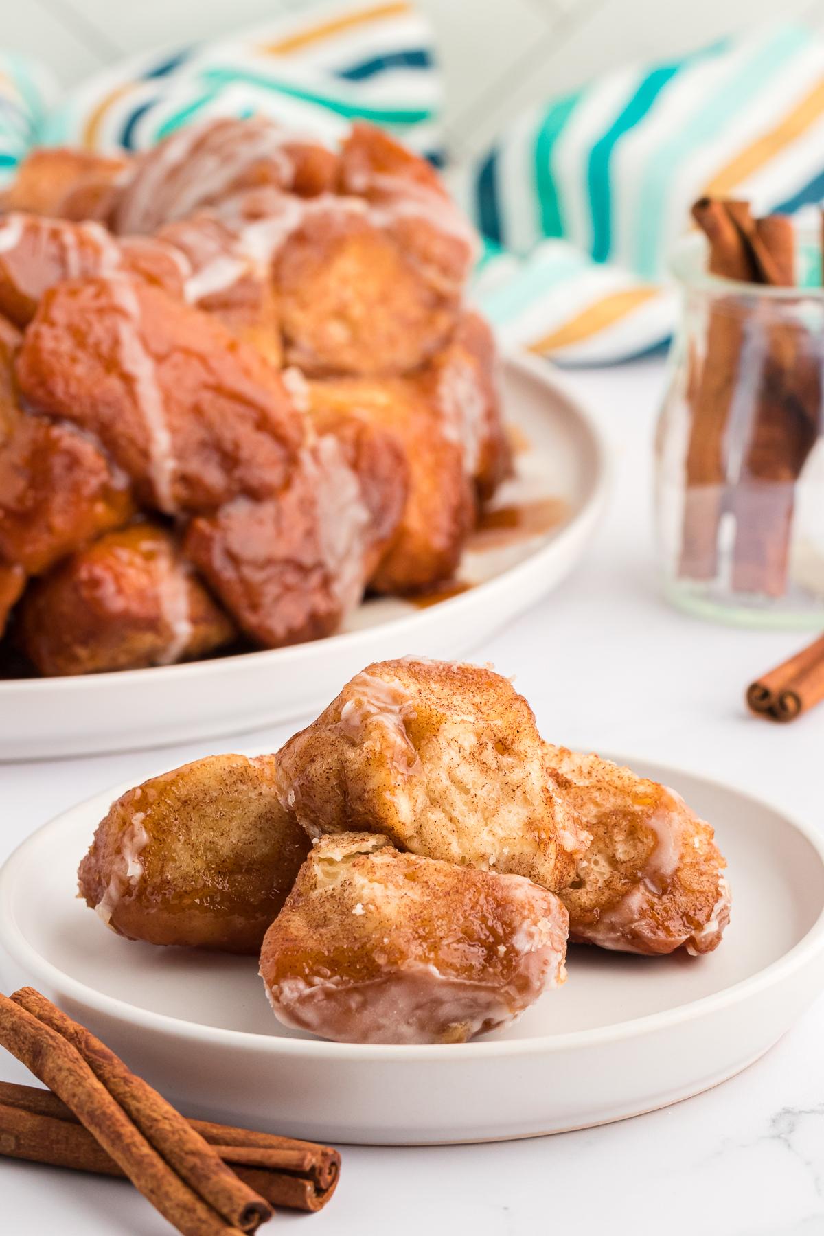 Pane delle scimmie di biscotti 15 Alcuni pezzi di Monkey Bread su un piccolo piatto bianco con bastoncini di cannella, un telo colorato a strisce e un piatto pieno di Monkey Bread intorno