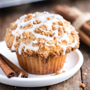 Muffin alla Sbriciolata 16 Un muffin al caffè con crumble sbriciolato e glassa bianca a filo è appoggiato su un piatto bianco, con due bastoncini di cannella accanto su una superficie di legno rustica.
