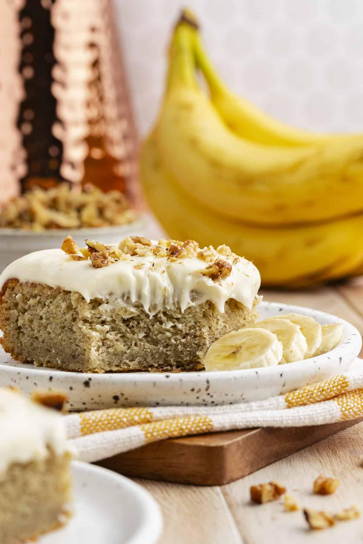 Ricetta Facile Torta alla Banana 16 Una fetta di torta di banane su un piccolo piatto su un canovaccio a strisce bianche e gialle su un tagliere di legno, circondata da altra torta, una ciotola di noci e banane intere