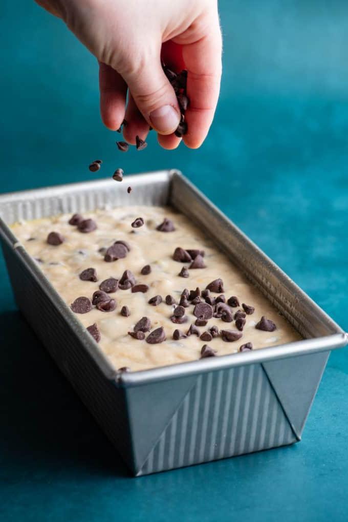 Il Miglior Pan di Banane con Gocce di Cioccolato 16 Impasto del pane alle banane e gocce di cioccolato in uno stampo da plumcake d'argento che viene cosparso di altre gocce di cioccolato, appoggiato su una superficie color ottanio scuro.