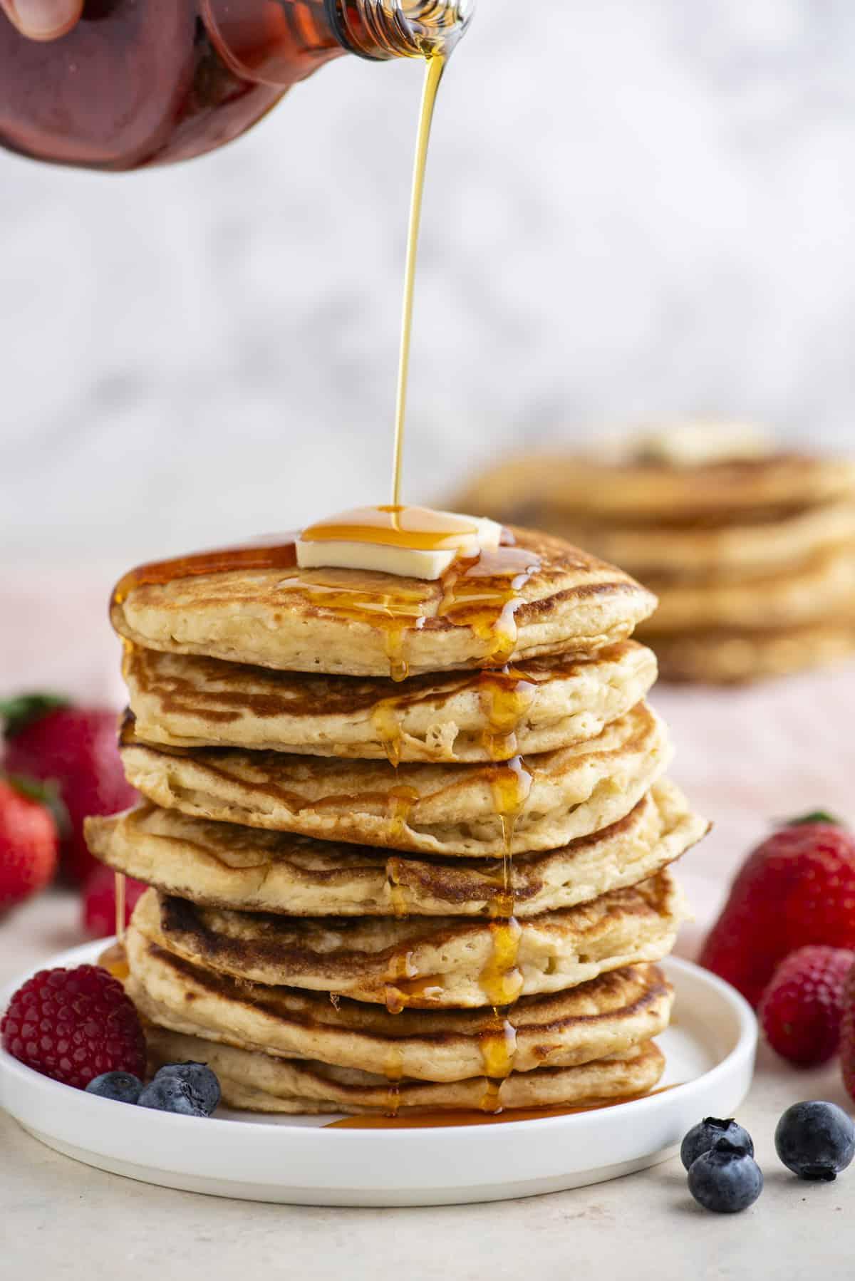 Pancakes Soffici Fatti in Casa 16 Una torre alta di pancake su un piatto bianco, condita con sciroppo, sormontata da una fetta di burro e circondata da frutti di bosco freschi, con altri pancake sullo sfondo.