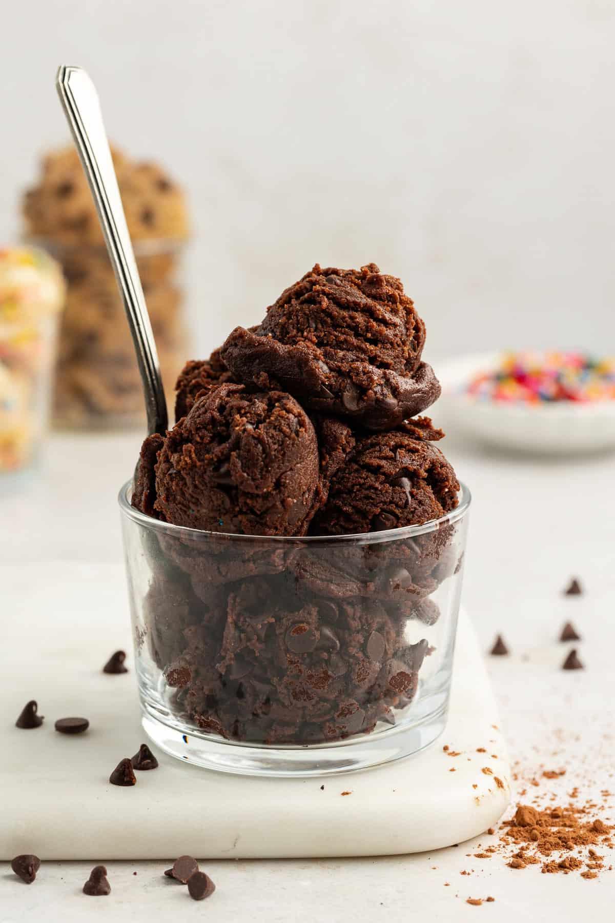 **Semplice Impasto per Biscotti Commestibile** 17 edible chocolate cookie dough balls in a clear glass container with a spoon on top of a white cutting board with chocolate chips sprinkled around and more cookie dough flavors in the background in clear glass containers