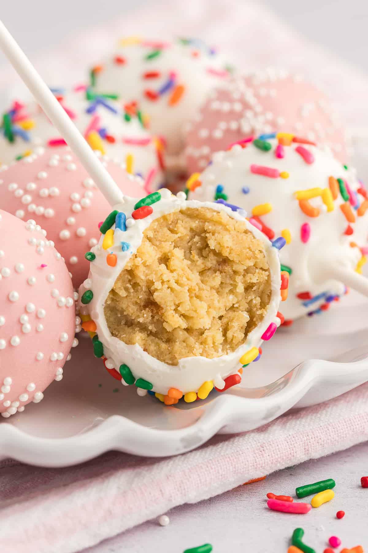 un piccolo piatto bianco su un asciugamano rosa con cake pops adagiati, con quello centrale anteriore a cui manca un morso che espone l'interno del cake pop