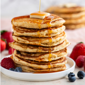Pancakes Soffici Fatti in Casa 17 Una torre alta di pancake su un piatto bianco, condita con sciroppo, sormontata da una fetta di burro e circondata da frutti di bosco freschi, con altri pancake sullo sfondo.