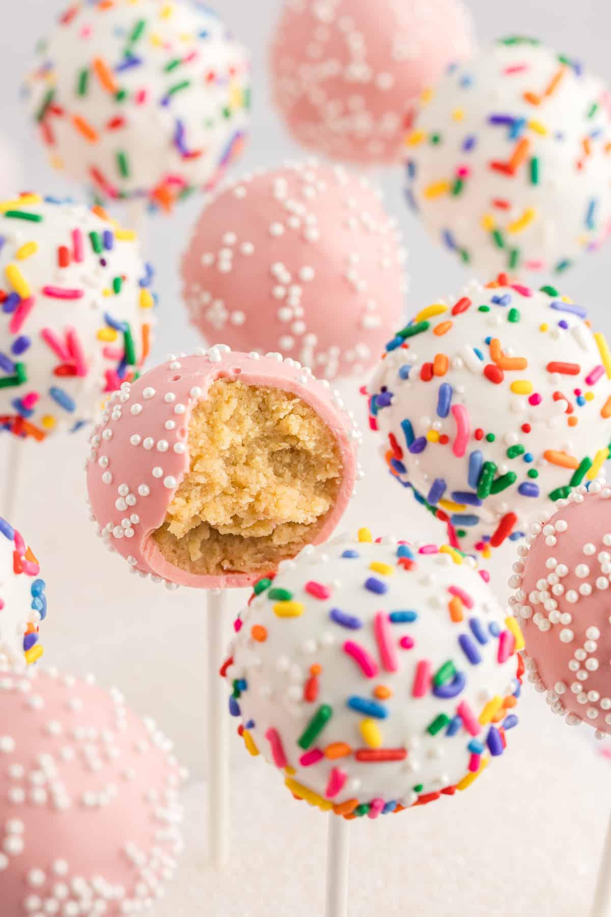 cake pops in piedi in un blocco di polistirolo bianco, alcuni rosa con codette bianche e alcuni bianchi con codette arcobaleno, con un cake pop rosa a cui manca un morso