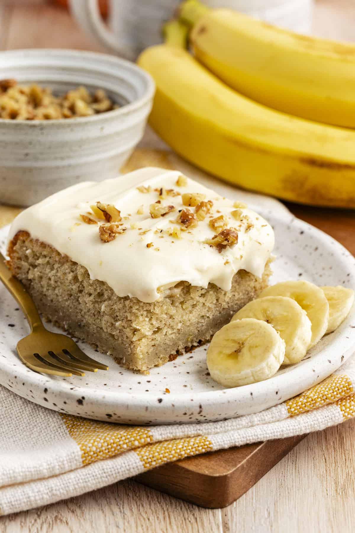 Ricetta Facile Torta alla Banana 18 Una fetta di torta di banane con un morso mancante su un piccolo piatto con fette di banana fresca, su un canovaccio da cucina a strisce gialle e bianche con banane intere e una ciotola di crumble in sottofondo
