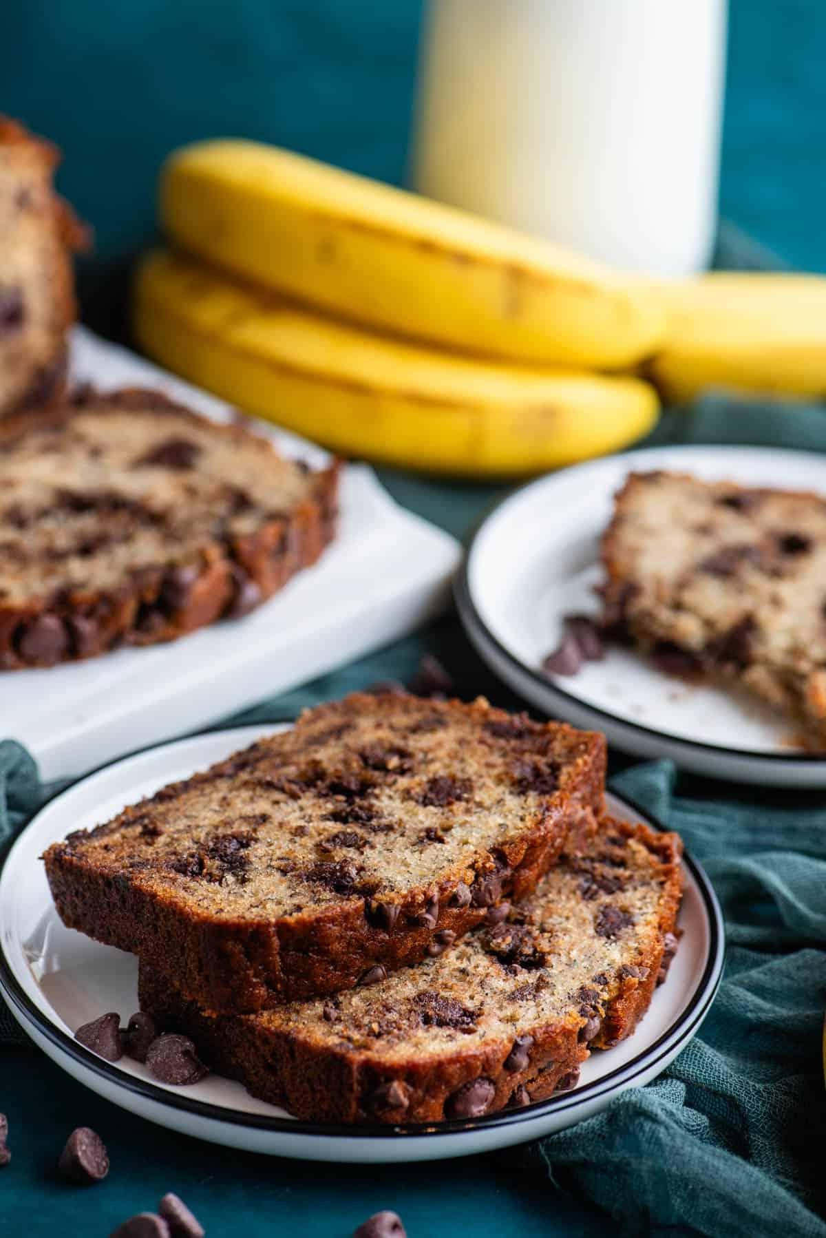 Il Miglior Pan di Banane con Gocce di Cioccolato 18 Due fette di pane alle banane e gocce di cioccolato su un piatto bianco sopra un asciugamano color ottanio scuro, con banane intere accanto, un altro piatto con una fetta di pane alle banane, e un tagliere bianco con altro pane dietro.