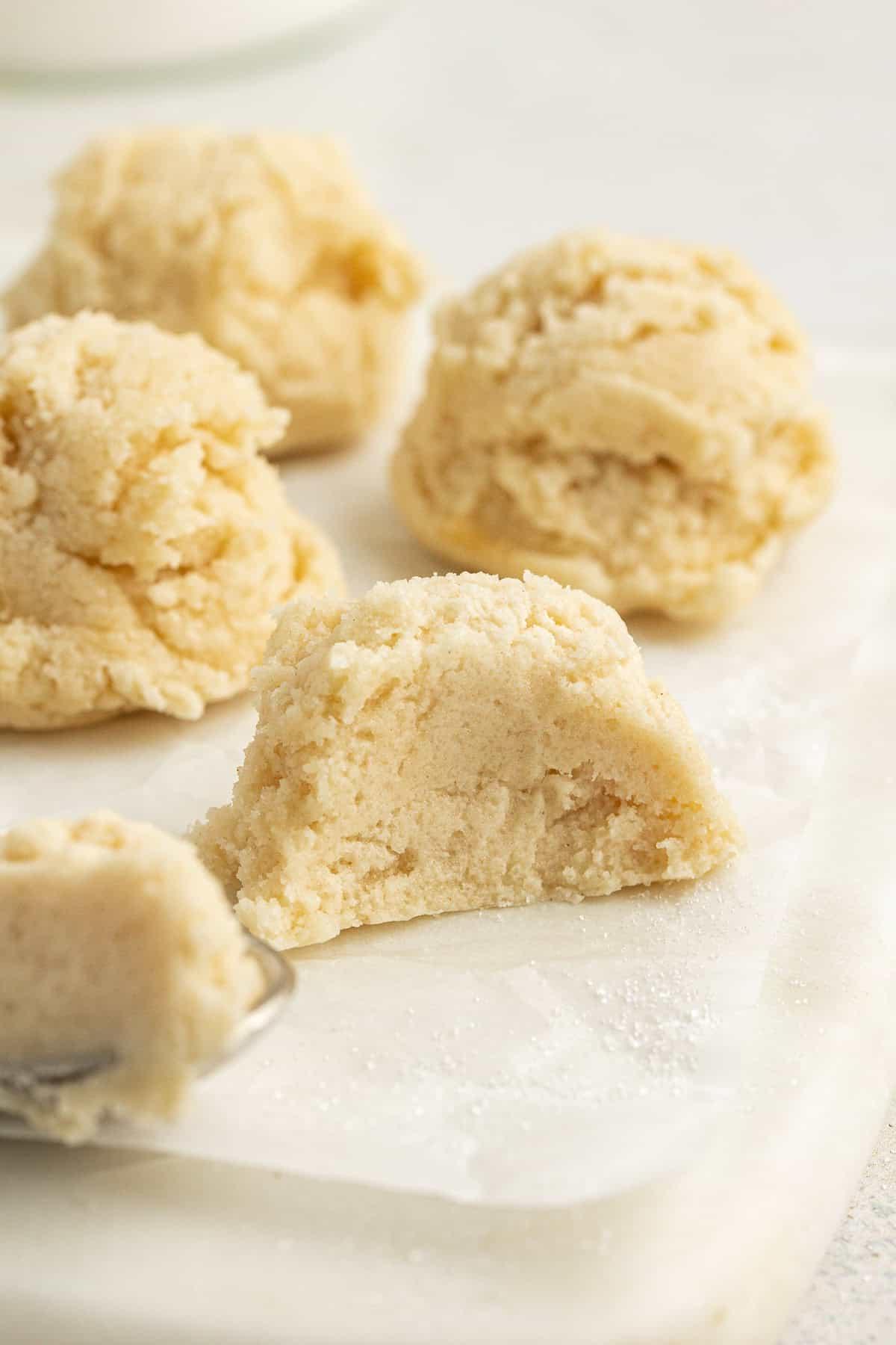 **Semplice Impasto per Biscotti Commestibile** 18 edible sugar cookie dough balls on a white cutting board with parchment paper with one ball sliced in half by a spoon and the spoon leaning on the cutting board with cookie dough on it