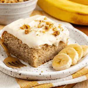Ricetta Facile Torta alla Banana 19 Una fetta di torta di banane con un morso mancante su un piccolo piatto con fette di banana fresca, su un canovaccio da cucina a strisce gialle e bianche con banane intere e una ciotola di crumble in sottofondo