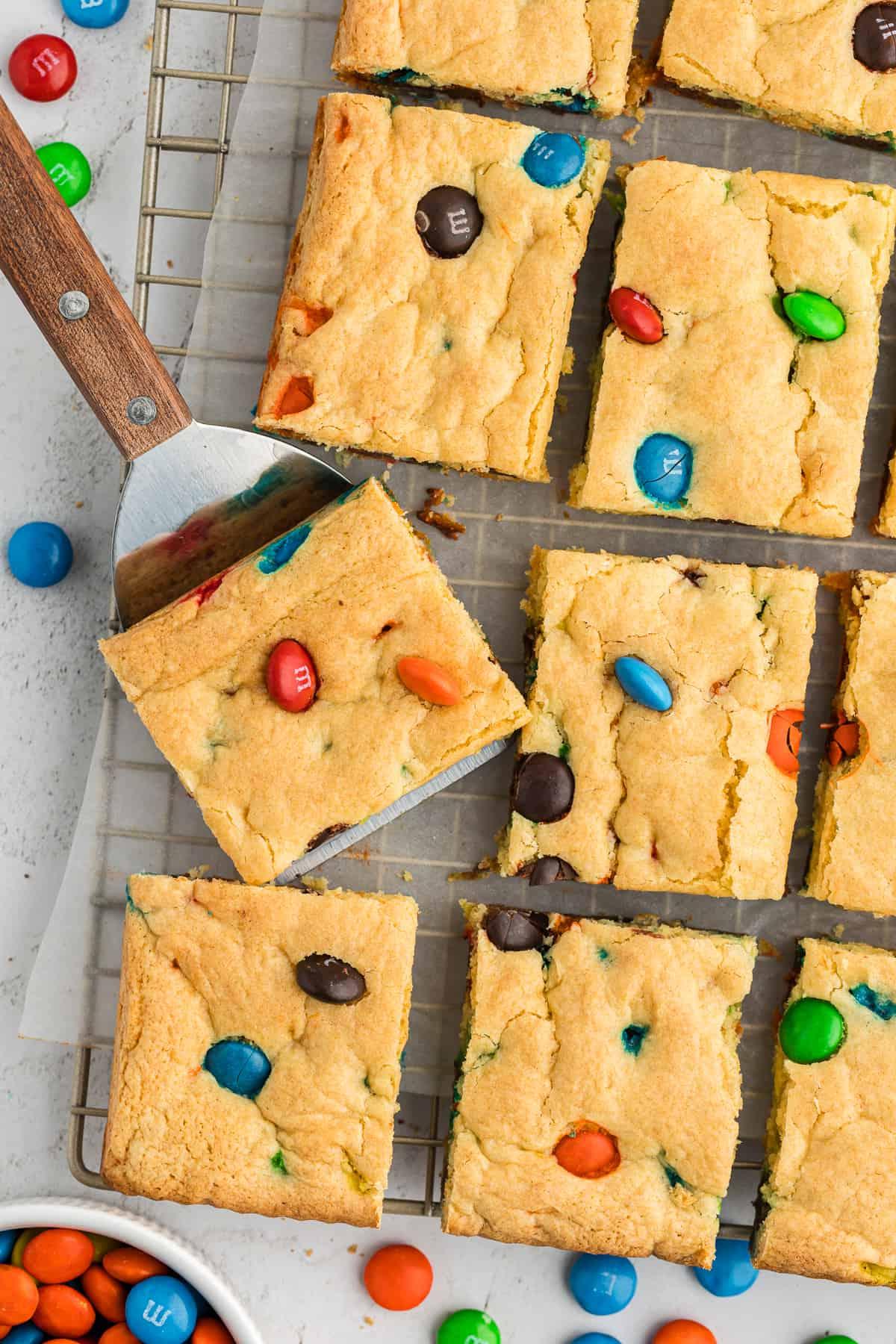 Barrette di biscotti da preparato per torte 19 rows of M&M cake bars on a wire rack, with one slightly lifted by a spatula, parchment paper under the wire rack and m&ms scattered around