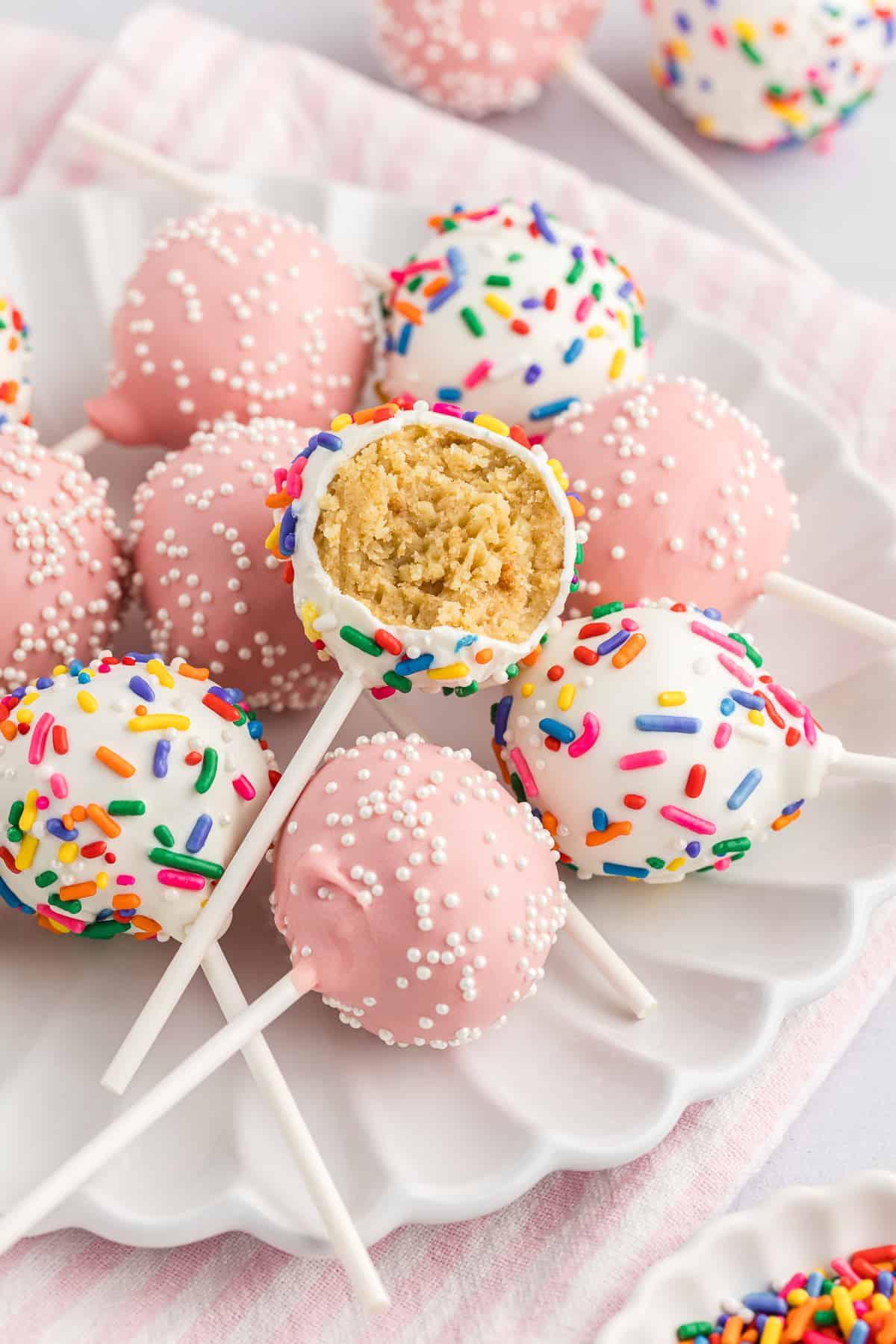 un piatto bianco con una pila di cake pops, alcuni rosa chiaro con codette bianche e alcuni bianchi con codette arcobaleno, con il cake pop in cima a cui manca un morso