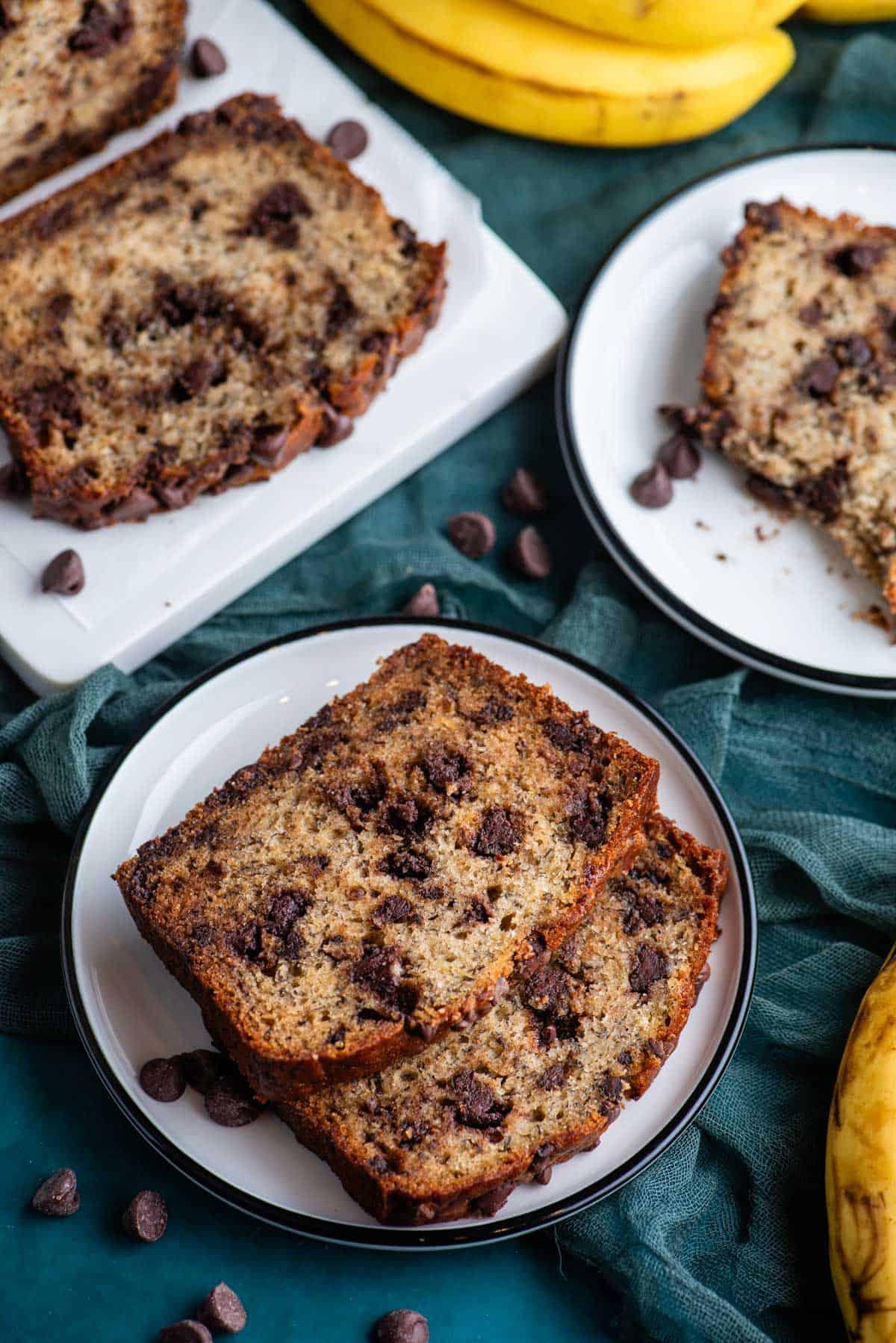 Il Miglior Pan di Banane con Gocce di Cioccolato 19 Due piccoli piatti bianchi con fette di pane alle banane, quello anteriore con due fette, e un tagliere bianco con fette di pane alle banane, il tutto appoggiato su un asciugamano color ottanio scuro con gocce di cioccolato sparse intorno.