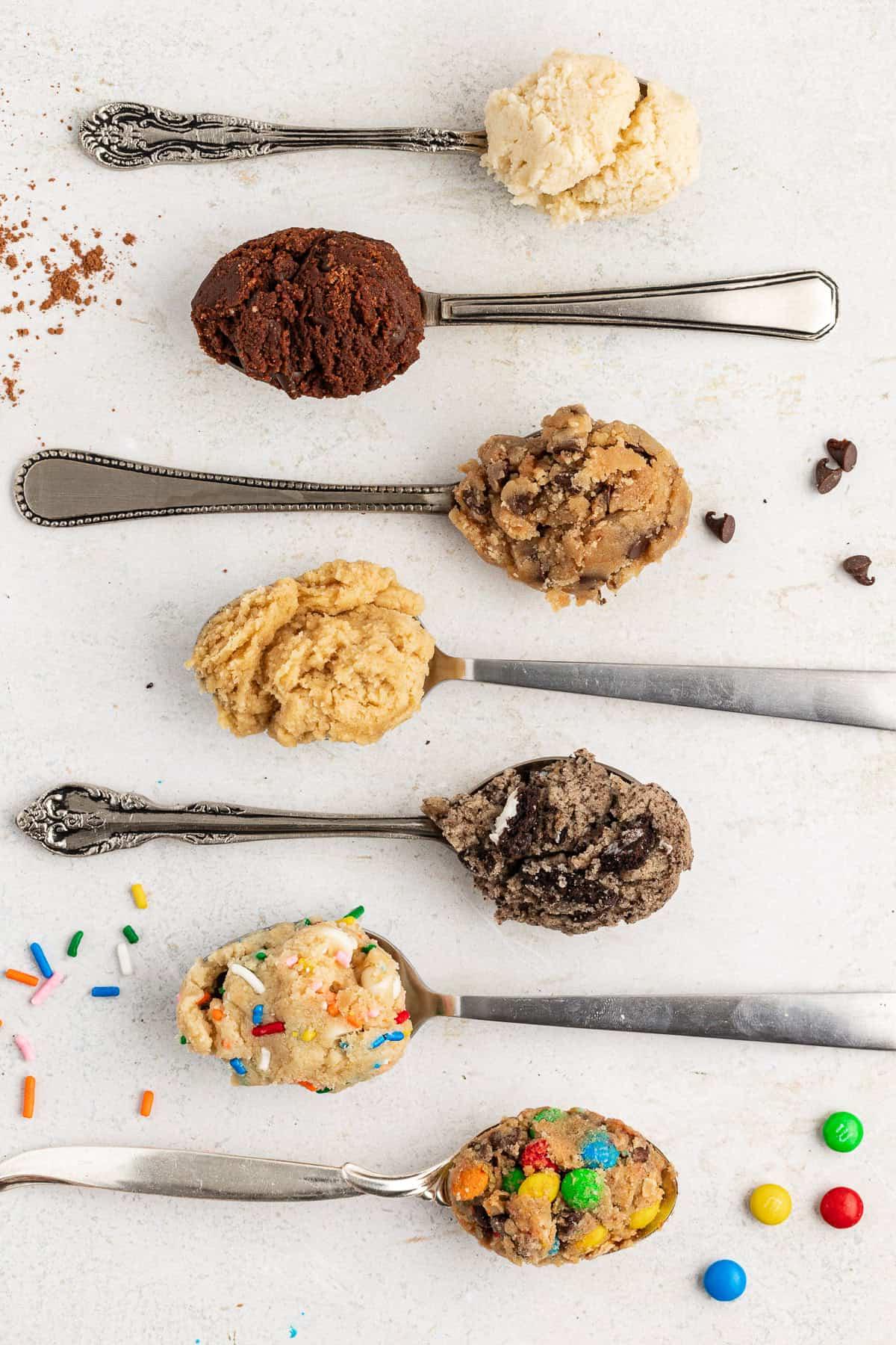 **Semplice Impasto per Biscotti Commestibile** 19 seven spoons alternating directions laying flat with edible cookie dough in them starting with sugar cookie, then chocolate, chocolate chip, peanut butter, oreo, funfettie and cookie monster at the bottom, with m&ms, sprinkles, chocolate chips, and cocoa powder sprinkled around