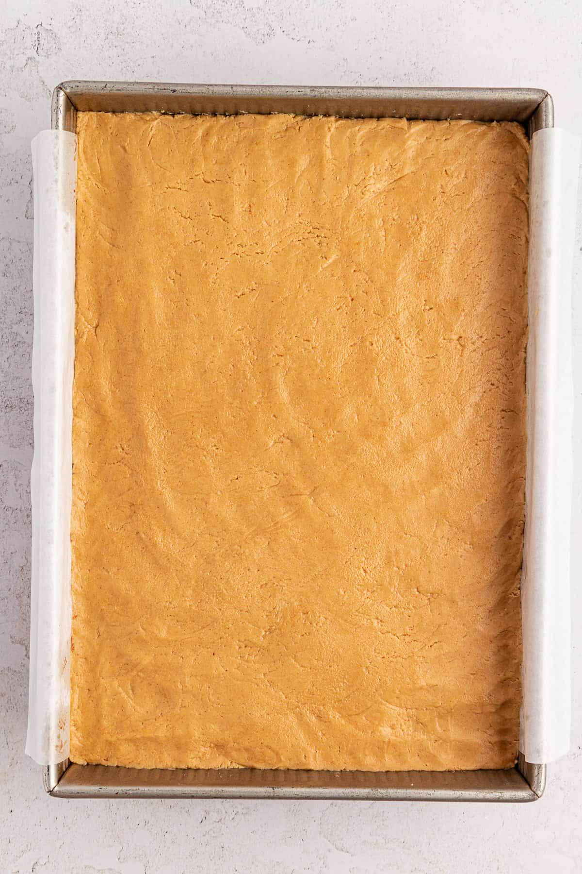 Una teglia da forno metallica quadrata foderata con carta forno, riempita con uno strato uniforme di miscela di burro d'arachidi sopra i brownies. L'impasto è liscio e compresso, pronto per la cottura.