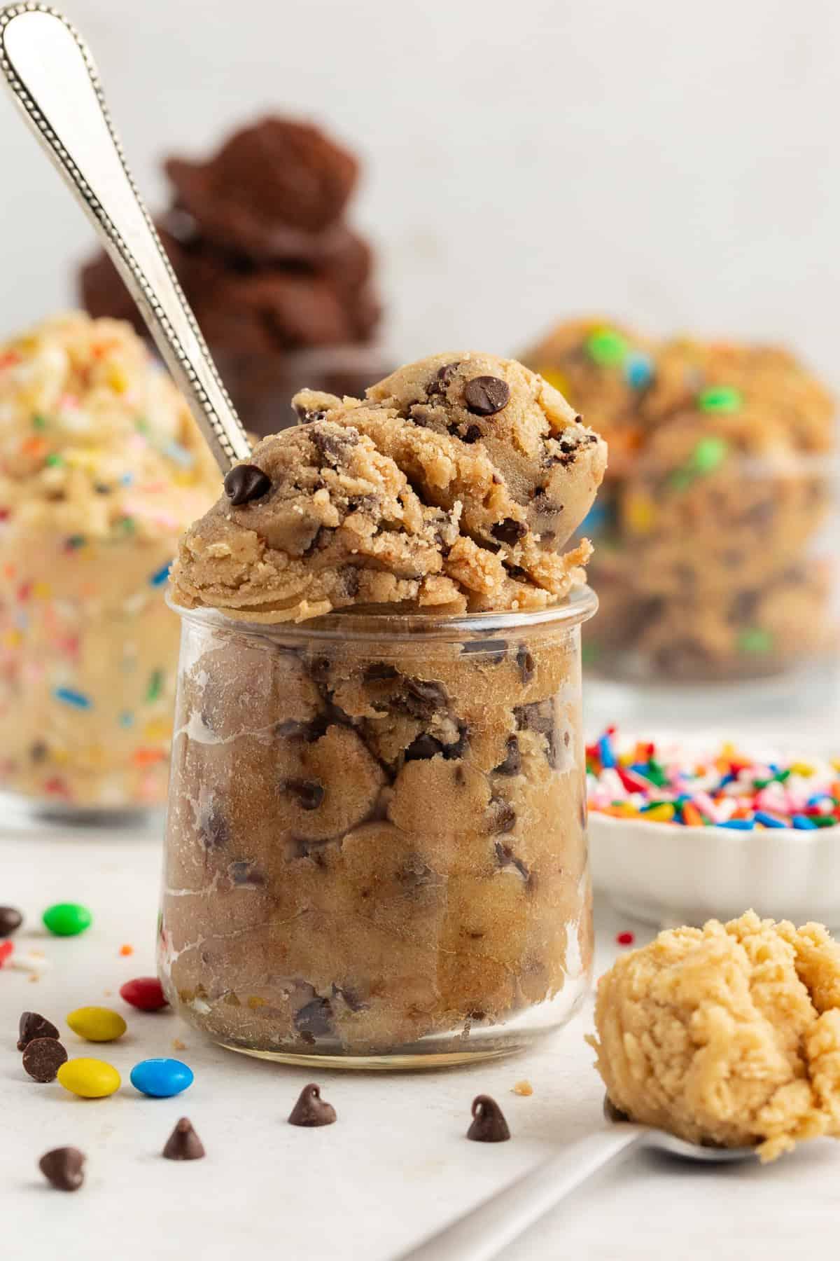 **Semplice Impasto per Biscotti Commestibile** 20 a clear glass jar filled with chocolate chip cookie dough with a spoon in it with m&ms and chocolate chips scattered around, more flavors of cookie dough in glass containers in the background and spoon full of peanut butter cookie dough in the front