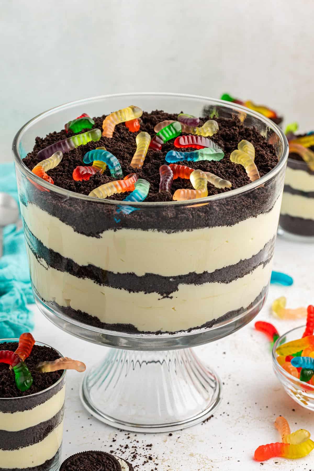 La migliore ricetta per la Torta al Fango 3 Una grande coppa di vetro trasparente da trifle piena di torta di terra guarnita con vermi gommosi, circondata da coppe più piccole di torta di terra, un asciugamano color ottanio, vermi gommosi e Oreo