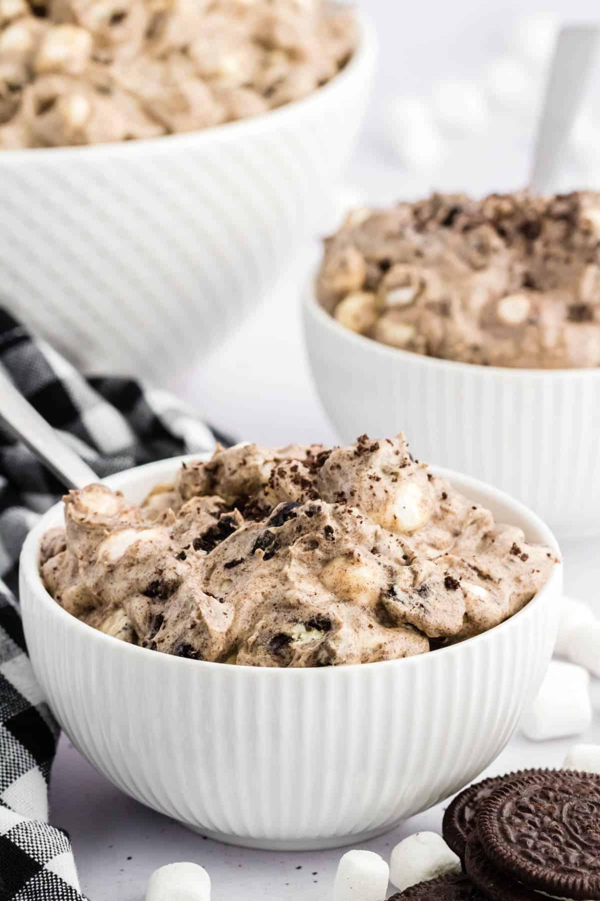 Ricetta Crema Soffice Oreo (Senza Formaggio Cremoso) 3 3 ciotole di fluff di Oreo circondate da biscotti Oreo interi, mini marshmallow e un panno da cucina a scacchi bianchi e neri