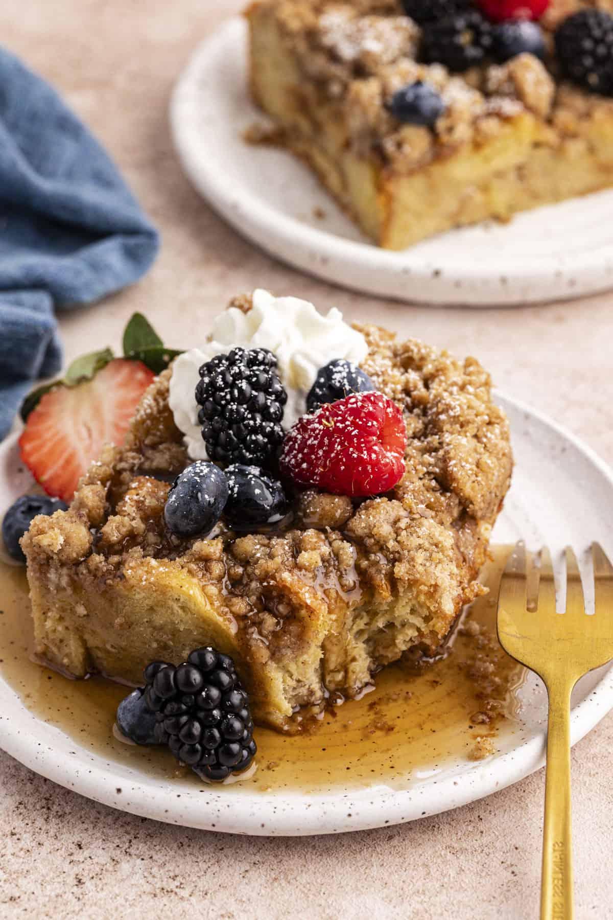 Migliore Ricetta per Budino di French Toast 3 Due piattini con una fetta di casseruola di French Toast ciascuno, coperti di sciroppo d'acero, frutti di bosco freschi, un ciuffo di panna montata e una leggera spolverata di zucchero a velo, con il piatto anteriore che ha una forchetta appoggiata e un canovaccio blu sullo sfondo.