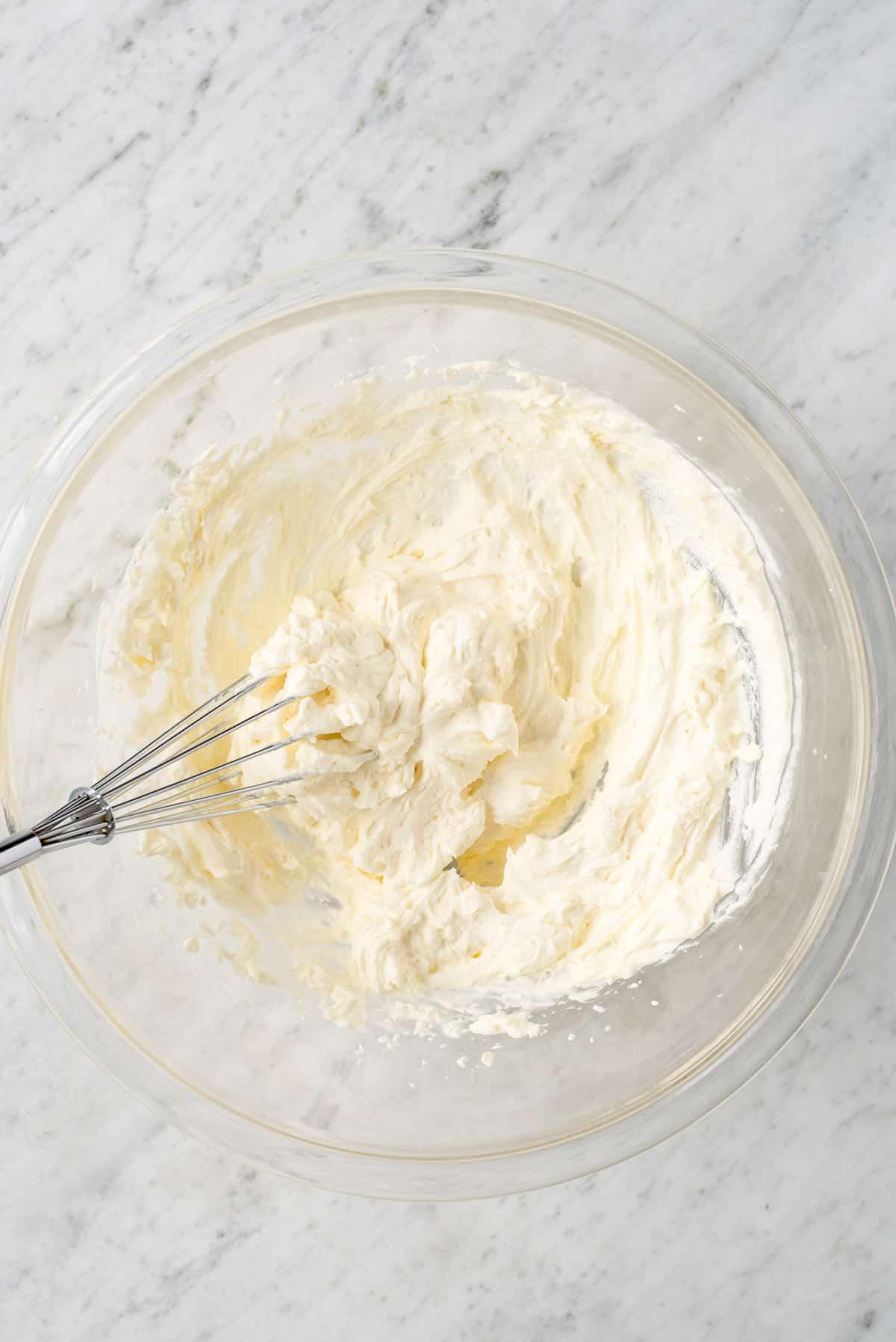 Come fare la panna montata al cheesecake stabilizzata che puoi usare su torte, cupcake, crostate e come salsa per frutta! Immagina questa panna montata al cheesecake su una crostata di fragole! #pannamonata #pannamonataaromatizzata #pannamonatacheesecake Panna montata al cheesecake in una ciotola di vetro con frusta