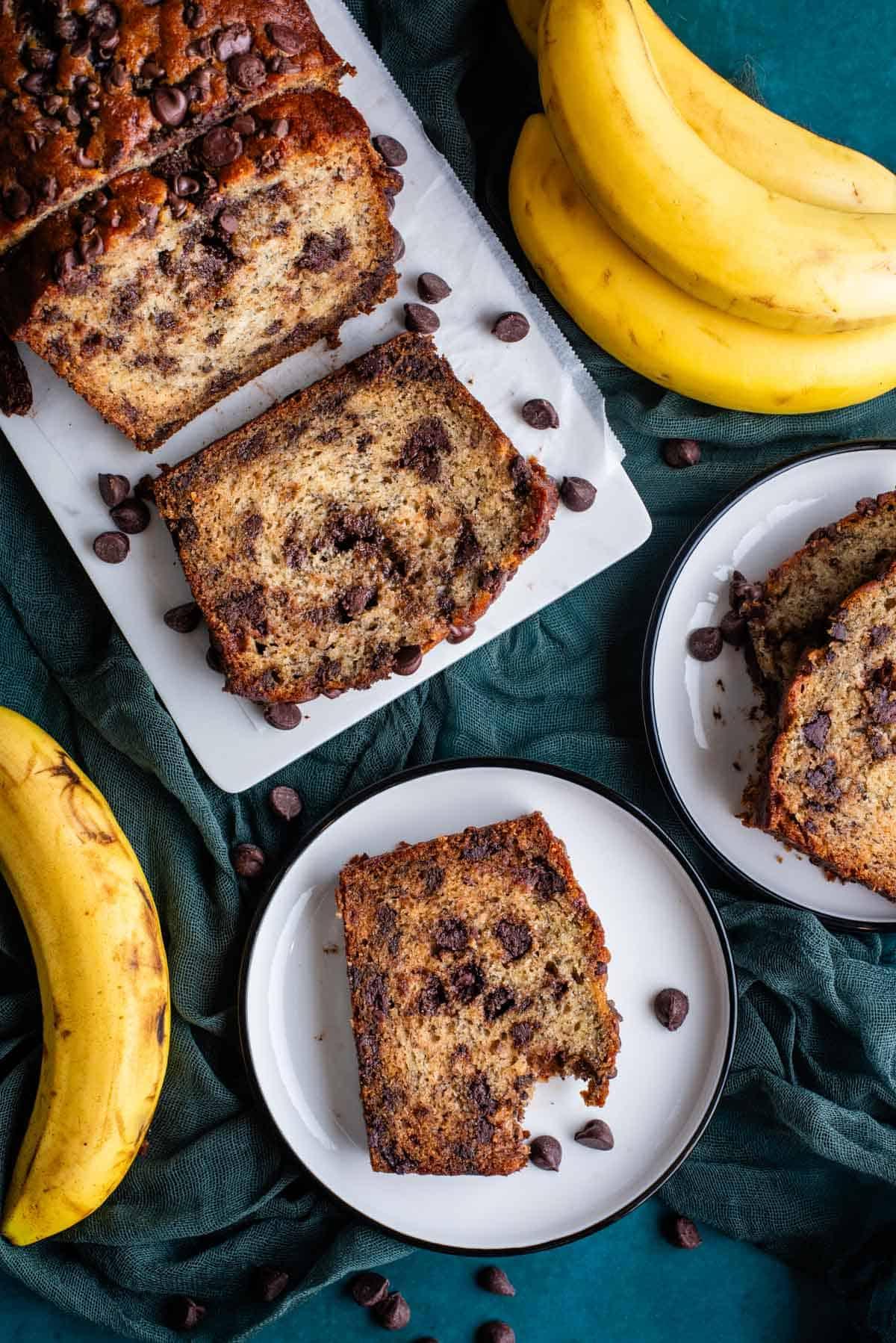 Il Miglior Pan di Banane con Gocce di Cioccolato 3 Vista dall'alto di due piatti bianchi e un tagliere bianco con fette di pane alle banane e gocce di cioccolato, tutti appoggiati su un asciugamano color ottanio scuro con gocce di cioccolato e banane intere sparse intorno.
