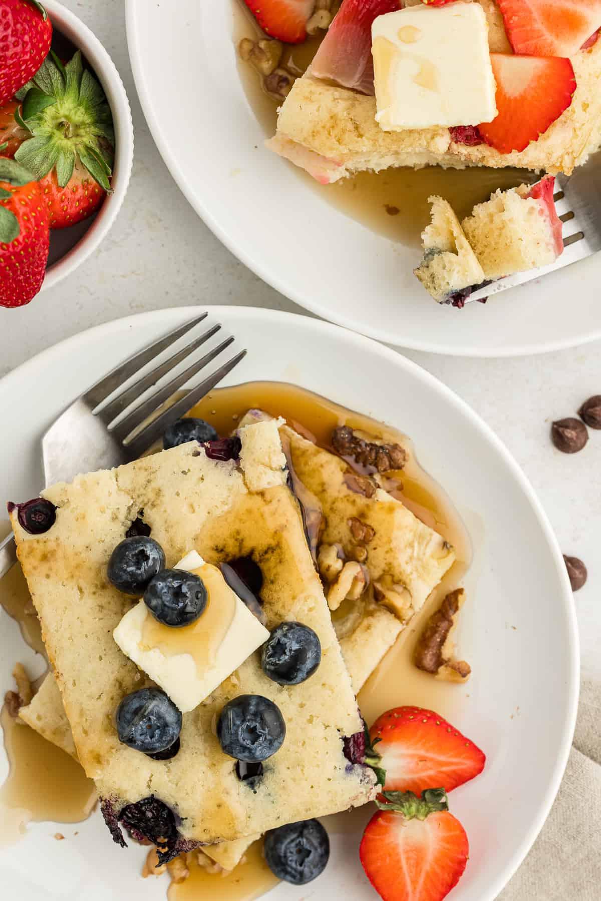 Ricetta Pancakes in Teglia 3 Due piatti bianchi con fette di pancake al forno in teglia, guarniti con fette di burro, sciroppo d'acero e mirtilli e fragole fresche, con forchette su entrambi i piatti e una ciotola di fragole fresche accanto.