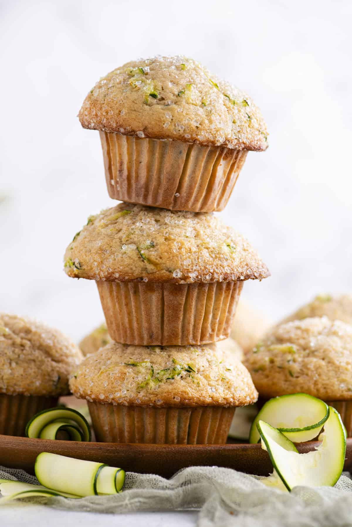 Migliori Muffin alle Zucchine 3 Una torre di tre muffin di zucchine uno sopra l'altro su un piatto di legno con altri muffin che li circondano e rotoli di sottili fette di zucchine sparse intorno
