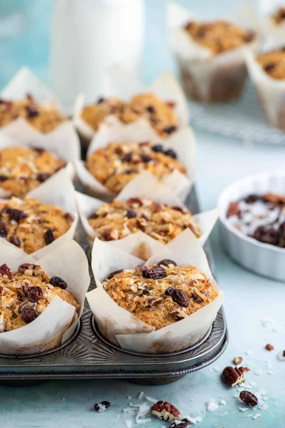 Muffin Morning Glory 3 Muffin Morning Glory nei pirottini di carta all'interno di una teglia per muffin, circondati da noci, uvetta e scaglie di cocco, con altri muffin sullo sfondo.