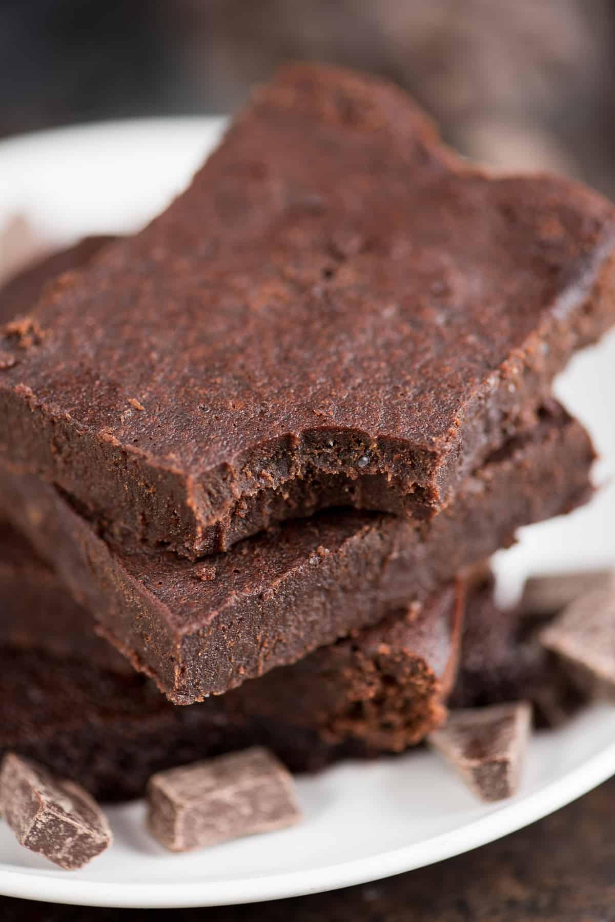 I brownies fondenti e salutari dei vostri sogni! Senza zucchero raffinato, burro o olio, con ingredienti comuni e un incredibile gusto fondente. Solo 174 calorie a fetta! Tre brownies fondenti salutari circondati da piccoli pezzi di cioccolato su un piatto da portata bianco.