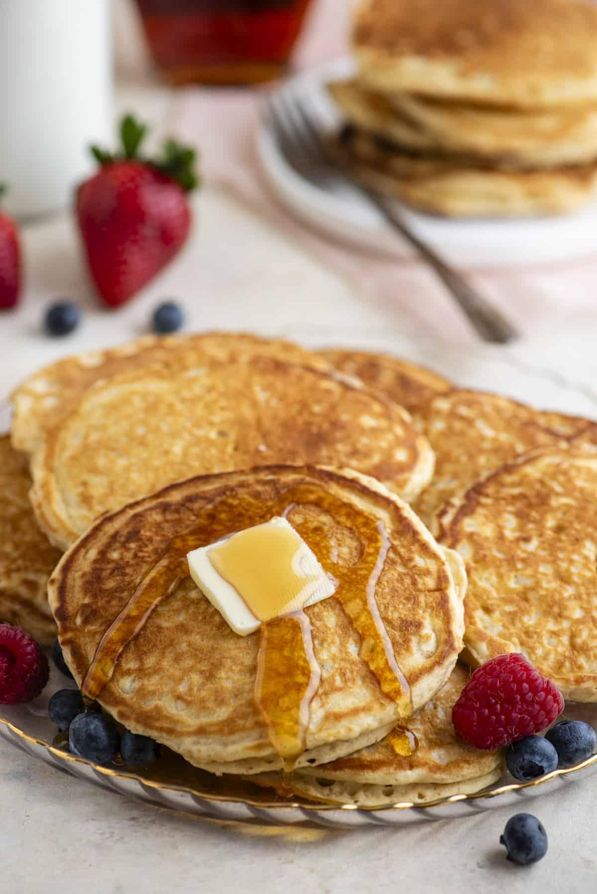 Pancakes Soffici Fatti in Casa 3 Pancake impilati su un piatto, uno dei quali sormontato da burro e condito con sciroppo, circondati da frutti di bosco freschi, con altri frutti di bosco e pancake sullo sfondo.