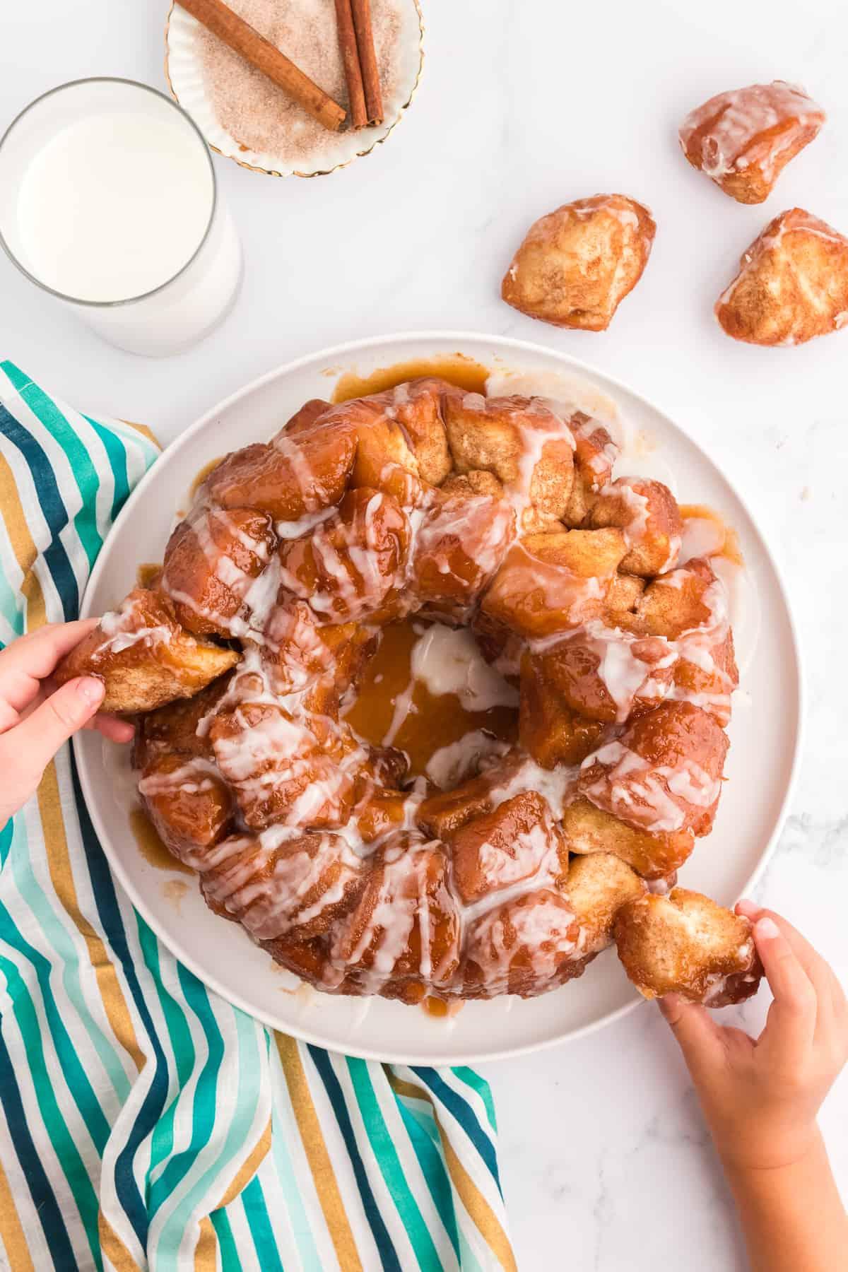 Pane delle scimmie di biscotti 3 Un piatto bianco con Monkey Bread e due piccole mani che staccano pezzi dal pane, un telo a strisce turchese, oro e blu sotto e pezzi di Monkey Bread, latte, zucchero alla cannella e bastoncini di cannella disposti intorno