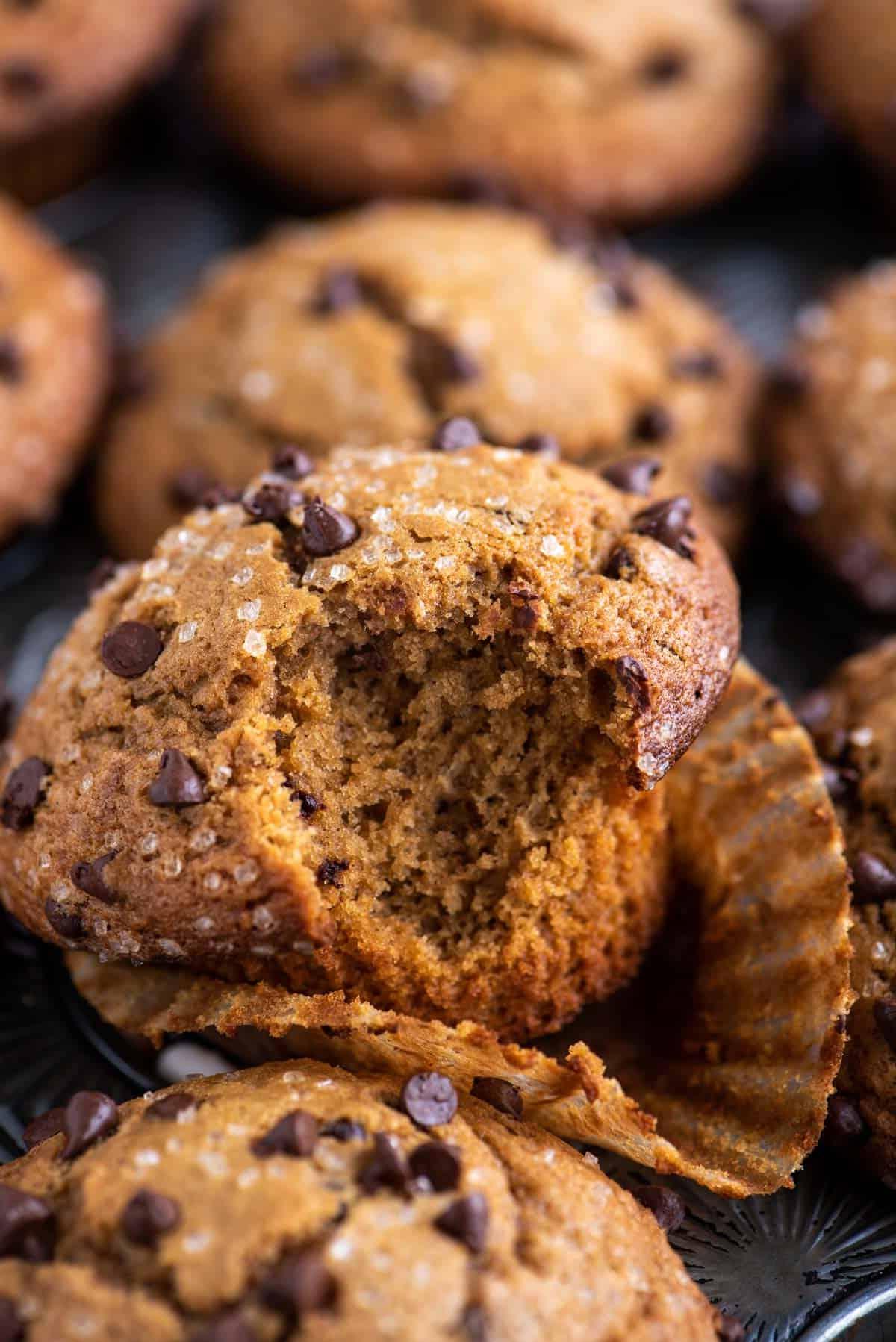 un muffin al cappuccino con gocce di cioccolato con un morso preso, appoggiato nel suo pirottino su una teglia per muffin piena di altri muffin