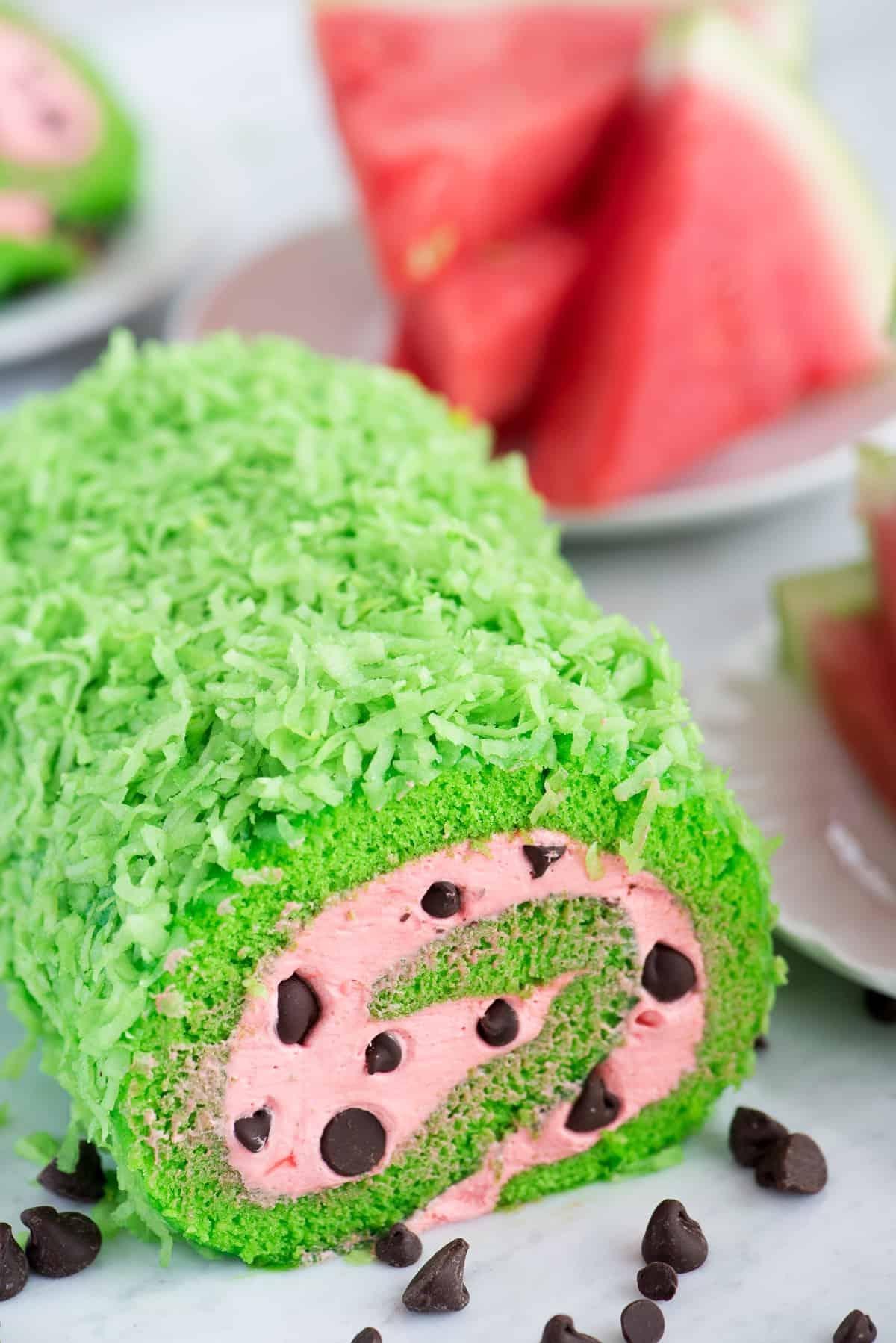Un rotolo di torta dall'aspetto fresco e verde con ripieno rosa e gocce di cioccolato, che richiama l'aspetto di una fetta di anguria.