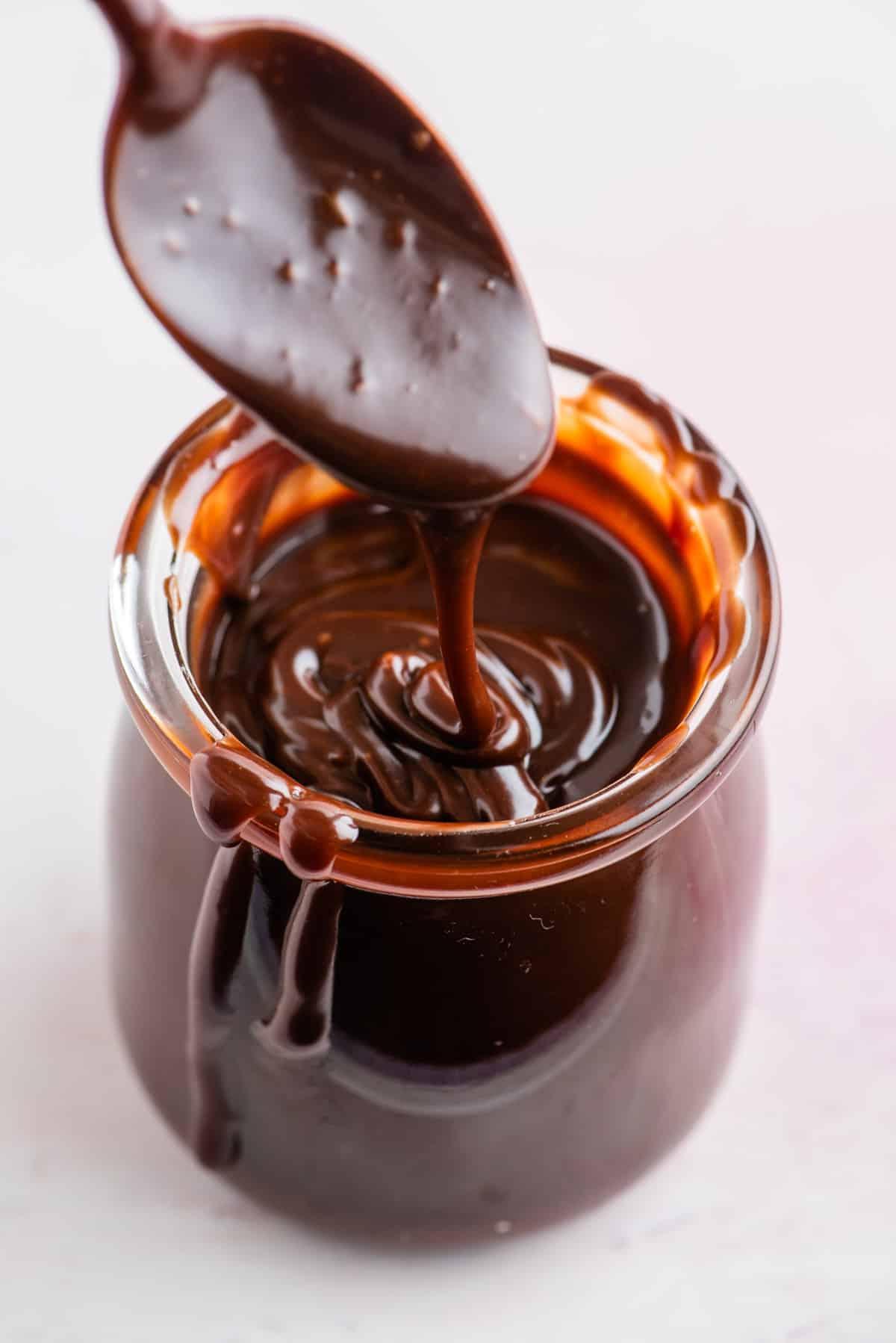 **Salsa Calda al Cioccolato Fondente** 3 Ricetta salsa hot fudge fatta in casa in un barattolo di vetro con un cucchiaio sopra il barattolo che gocciola un po' di salsa nel barattolo