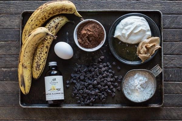 Questa torta al cioccolato più sana sa di muffin alla banana con doppie gocce di cioccolato! Niente zucchero, burro o olio, ma usa banane, yogurt greco e miele invece! Vassoio con ingredienti per la cottura: banane, yogurt, burro di arachidi, farina, gocce di cioccolato, uovo