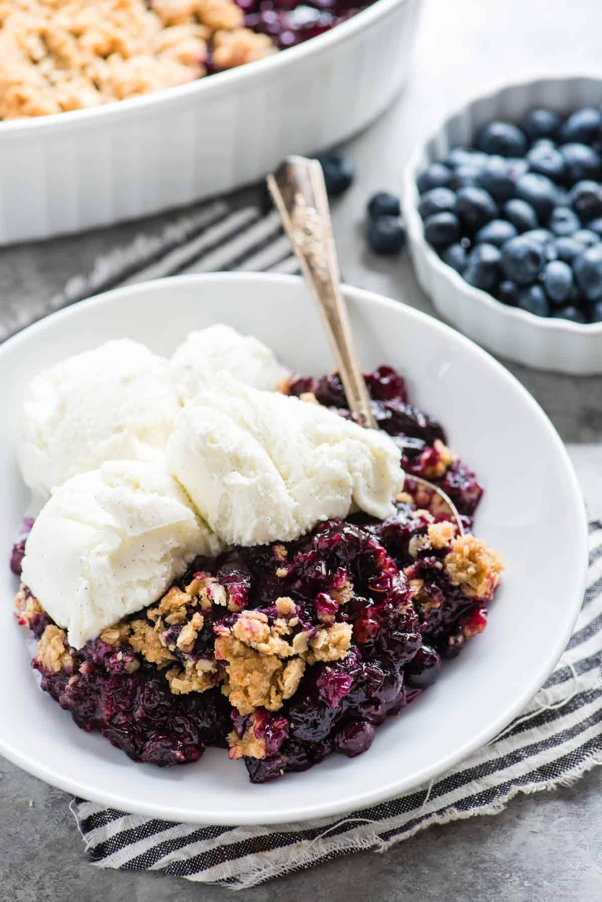 Crumble di Mirtilli Freschi 3 Crumble ai mirtilli in una ciotola bianca con gelato alla vaniglia e un cucchiaio di metallo su un tovagliolo a righe blu