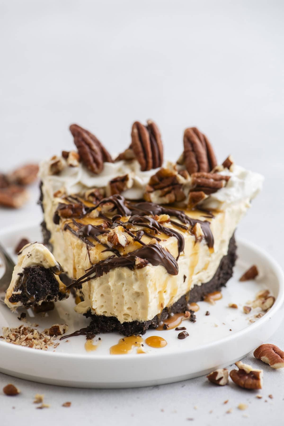 Torta di Tartaruga 3 Una fetta di Torta Tartaruga guarnita con noci pecan, gocce di cioccolato e caramello, su un piccolo piatto bianco con pezzi di noci pecan sparsi intorno.