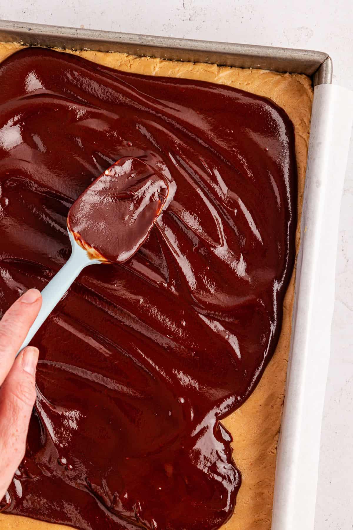 Una mano spalma glassa di cioccolato lucida su una teglia rettangolare di brownies Buckeye in una teglia da forno foderata con carta pergamena.
