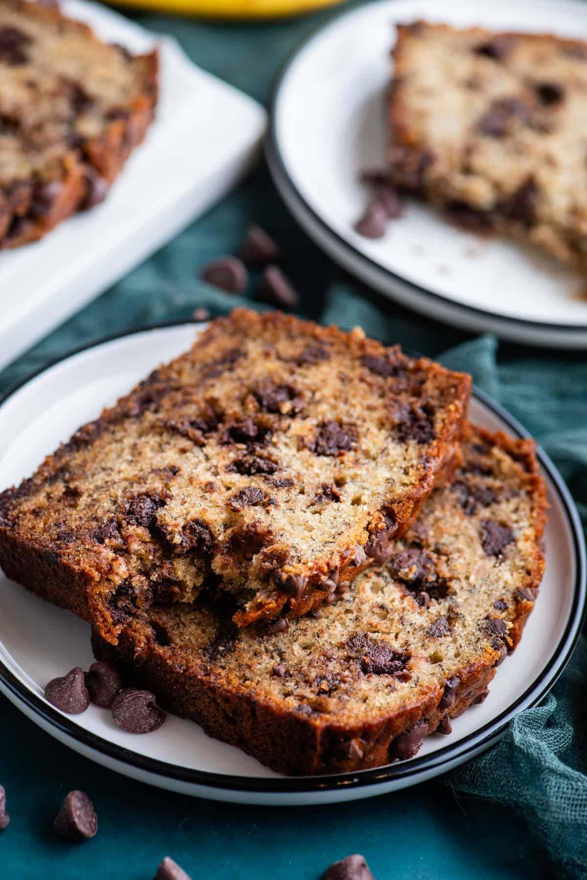 Il Miglior Pan di Banane con Gocce di Cioccolato 21 Due fette di pane alle banane e gocce di cioccolato su un piccolo piatto bianco su un asciugamano color ottanio scuro, con un morso preso dalla fetta superiore, un altro piatto con pane e un tagliere bianco con pane sullo sfondo e gocce di cioccolato sparse intorno.