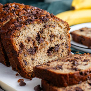 Il Miglior Pan di Banane con Gocce di Cioccolato 23 Pane alle banane e gocce di cioccolato affettato su un tagliere bianco con un piatto sullo sfondo con una fetta e banane intere dietro.