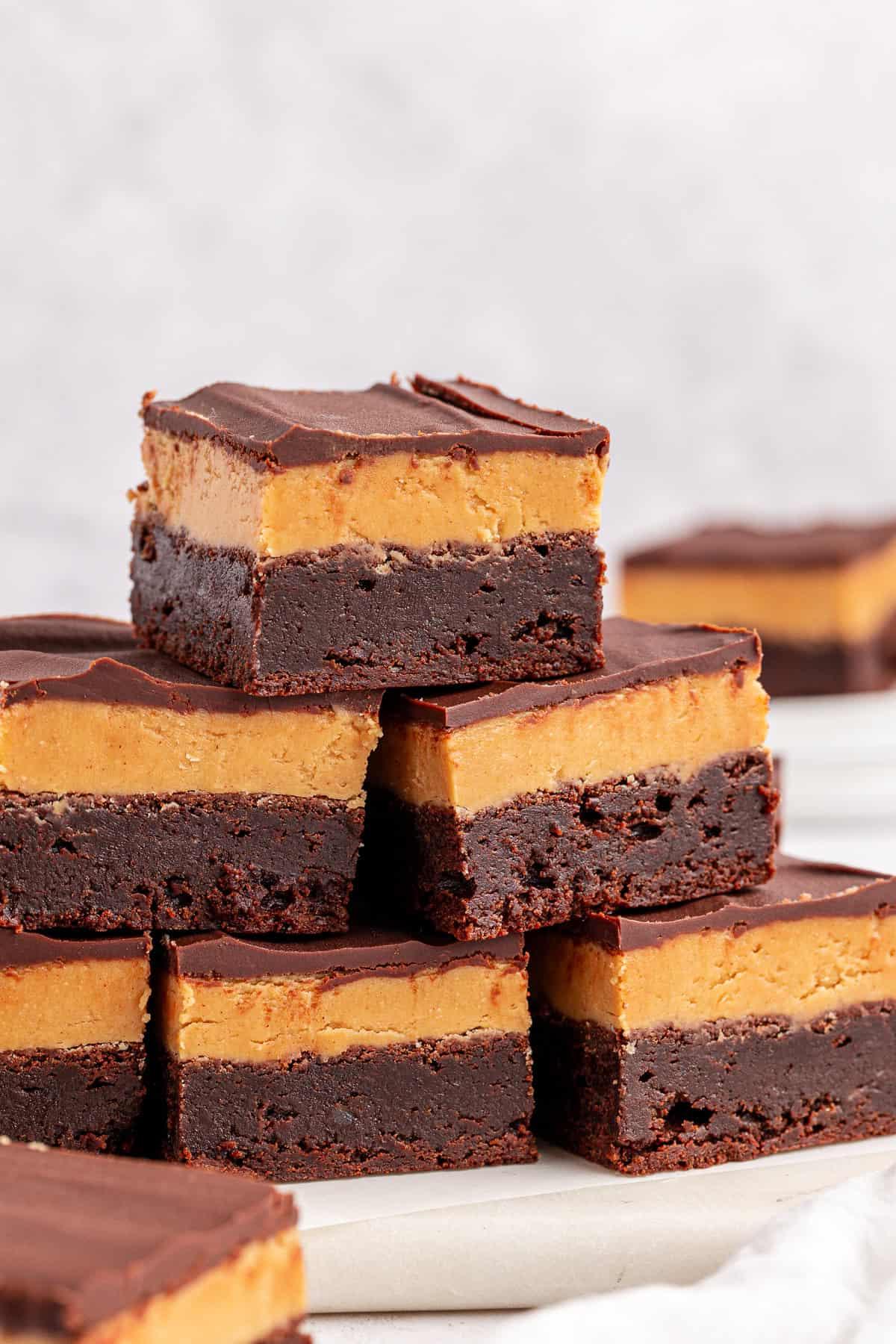 Una pila di ricchi brownies Buckeye con uno spesso strato di burro d'arachidi e una morbida copertura di cioccolato, disposti su una superficie bianca con altri brownies sfocati sullo sfondo.