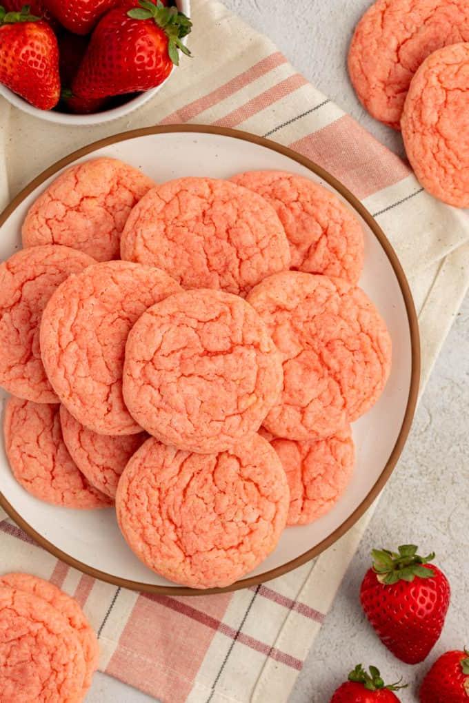 Fare Biscotti dal Mix per Torte 27 Biscotti alla fragola con preparato per torte impilati su un piatto bianco con bordo marrone, appoggiato su un canovaccio da cucina beige e rosa e circondato da altri biscotti, fragole e una piccola ciotola bianca di fragole