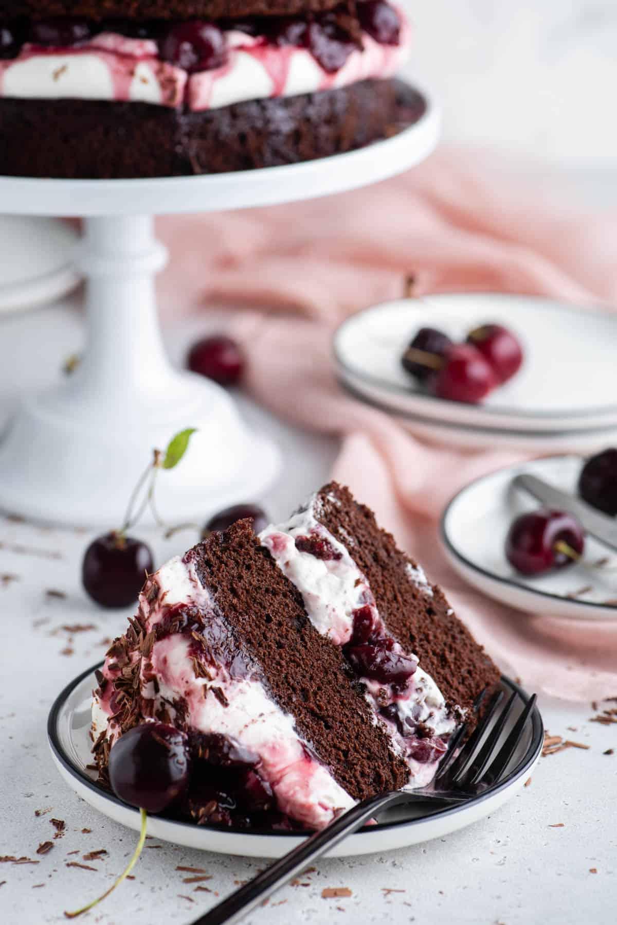 Torta Foresta Nera 4 Una fetta di Torta Foresta Nera su un piatto bianco con un'alzatina e altri piatti sullo sfondo e ciliegie e scaglie di cioccolato sparse intorno