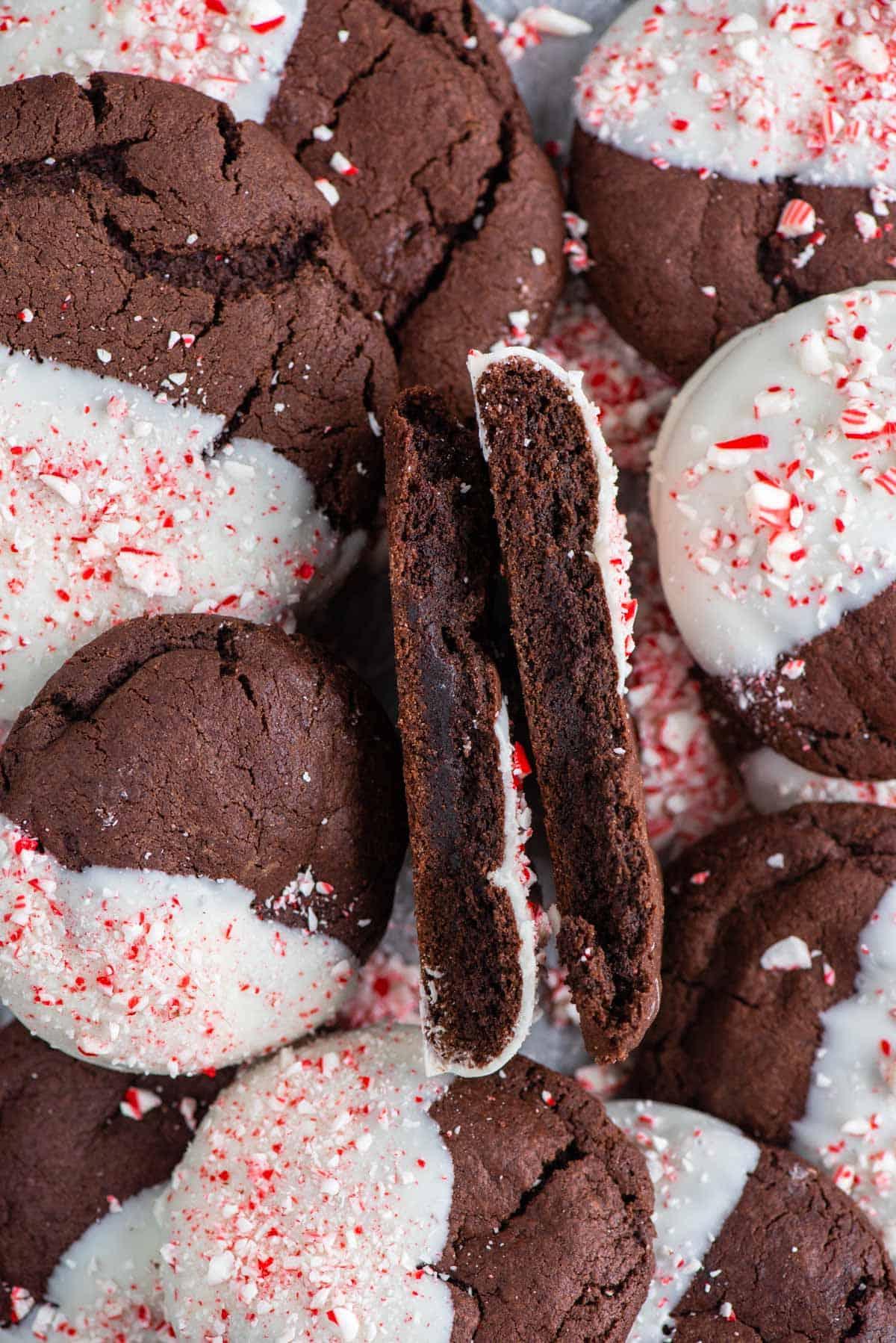 Questi Biscotti al Cioccolato e Bastoncini di Zucchero sono morbidi, gommosi, immersi nel cioccolato bianco e cosparsi di bastoncini di zucchero tritati. Un biscotto al cioccolato e bastoncini di zucchero tagliato a metà sopra altri biscotti