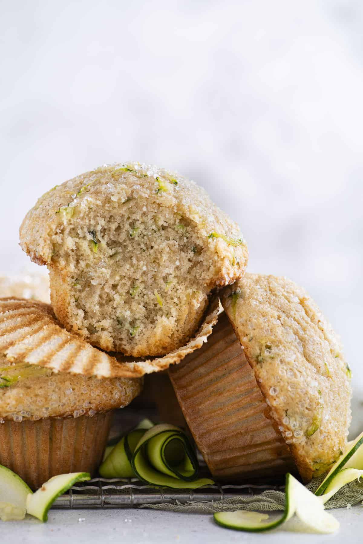 Migliori Muffin alle Zucchine 4 Un mucchio di tre muffin di zucchine su una griglia di raffreddamento con sottili fette di zucchine sparse intorno, e il muffin superiore con il suo pirottino rimosso e un morso mancante dal muffin